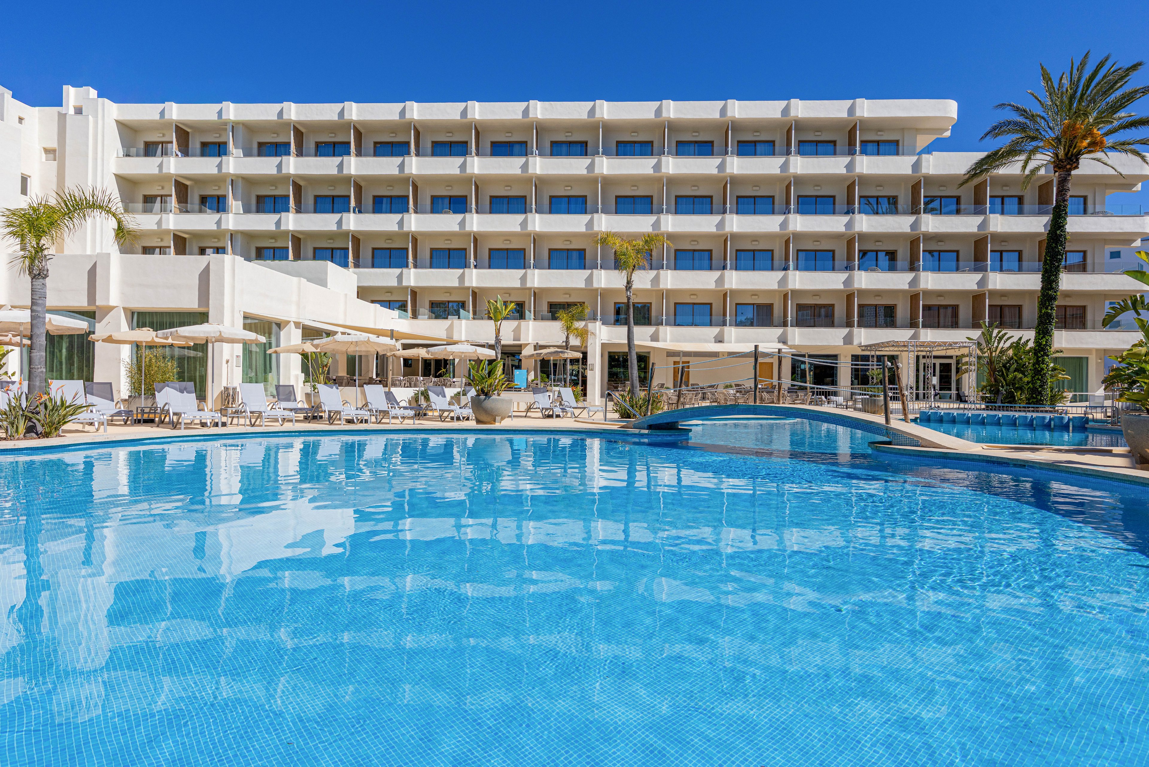 A large modern hotel building overlooks a bright blue outdoor swimming pool. The multi-story structure features rows of balconies facing the water, with sun loungers and umbrellas arranged around the pool deck. Clear blue sky and palm trees enhance the resort-like atmosphere. The scene suggests a sunny vacation destination focused on relaxation and leisure by the pool.