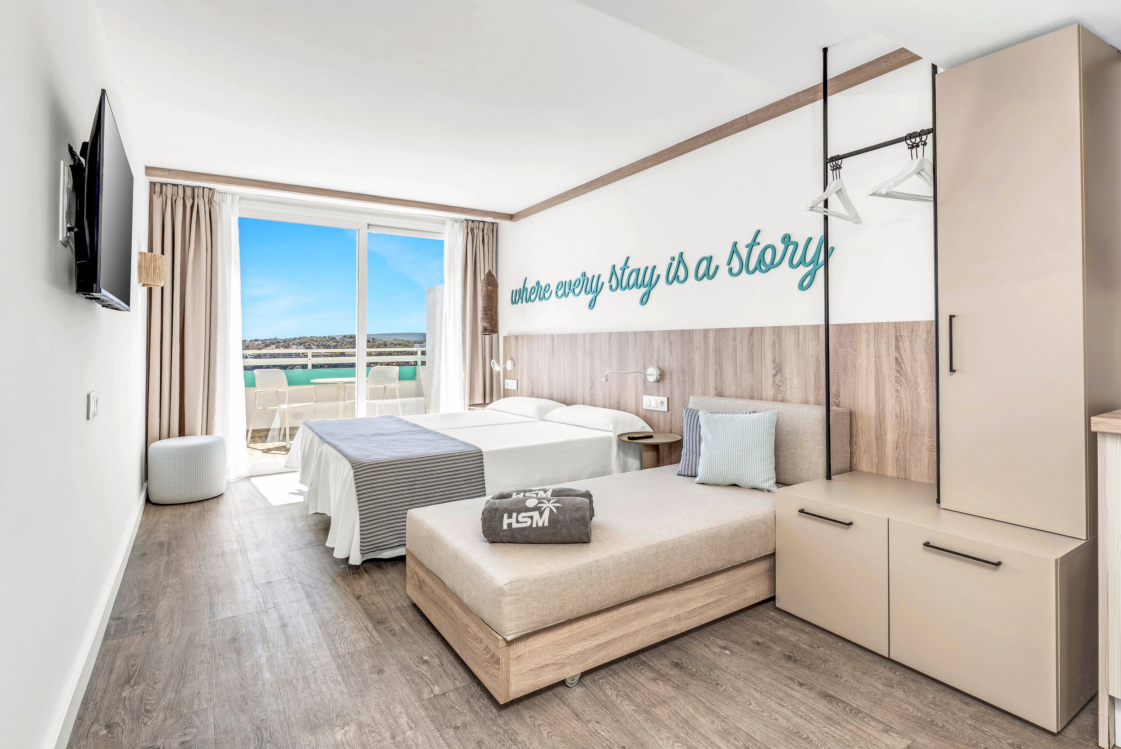 Modern hotel room featuring two beds and a daybed overlooks a sunny seaside balcony. Large sliding glass doors let in natural light and reveal blue sky and water outside. The interior decor is minimalist with light wood furniture, neutral textiles, and soft blue accents. A wall quote reading "where every stay is a story" adds a welcoming touch to the contemporary space.