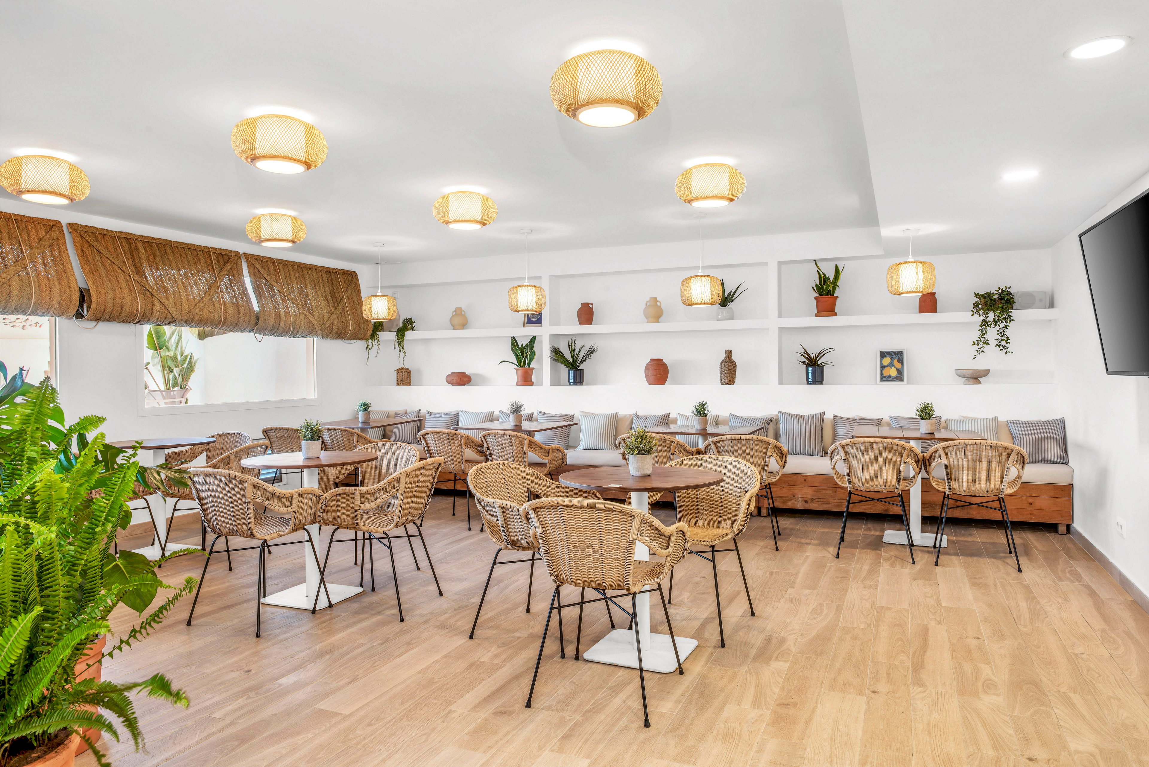 A bright, modern cafe interior features several small tables with wicker chairs arranged on a light wooden floor. Built-in bench seating with neutral cushions lines the back wall, creating a cozy dining area. White walls and shelves display potted plants and decorative vases, adding natural texture and color. Warm ceiling lights and large windows with woven shades enhance the airy, inviting atmosphere.