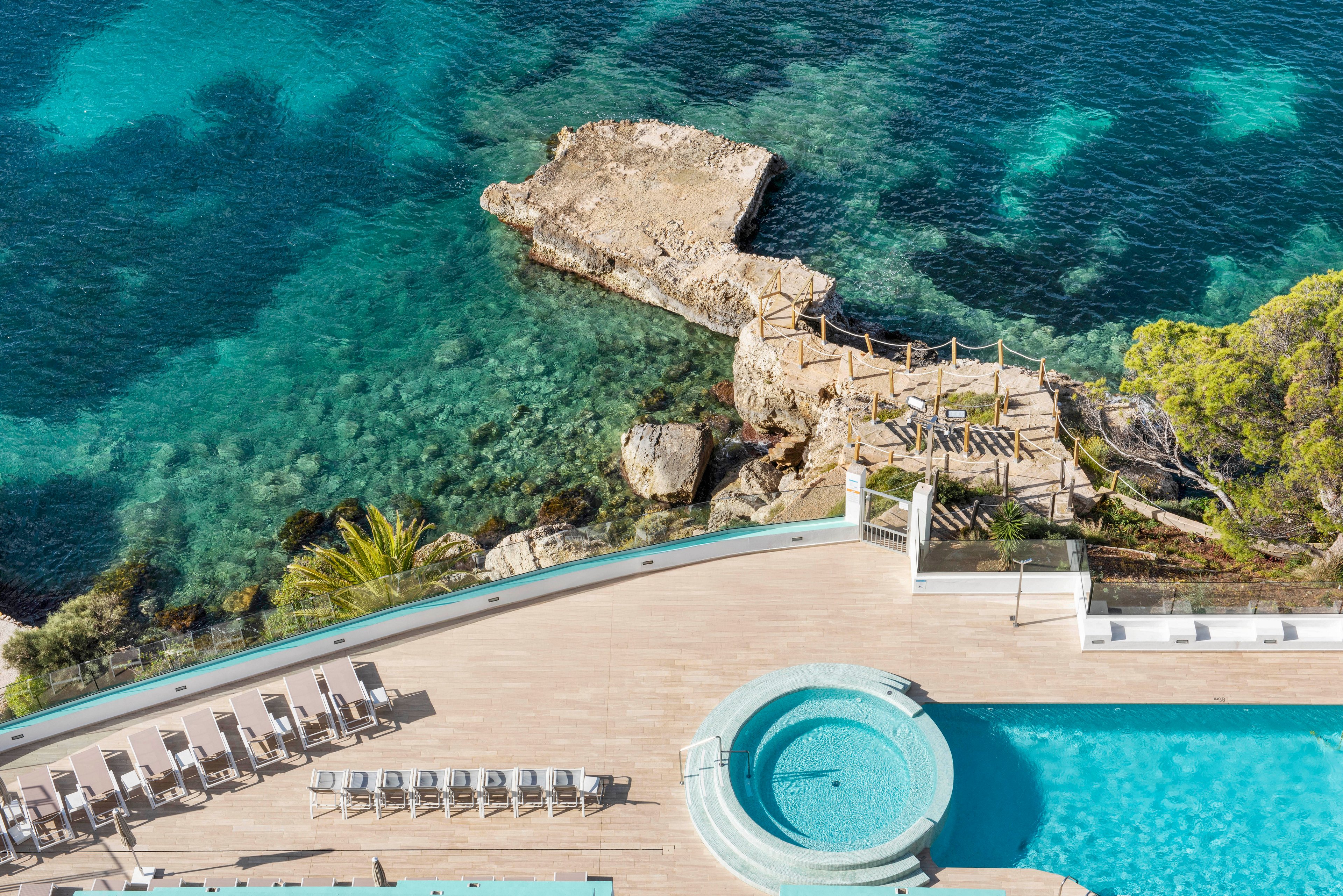 An elevated view shows a resort swimming pool and circular hot tub beside a wooden deck. Empty lounge chairs line the pool area, suggesting a quiet, off-peak moment. Beyond the terrace, clear turquoise seawater laps against a rocky shoreline with a small wooden stairway leading down. Lush greenery and the vivid blue ocean create a serene coastal vacation atmosphere.