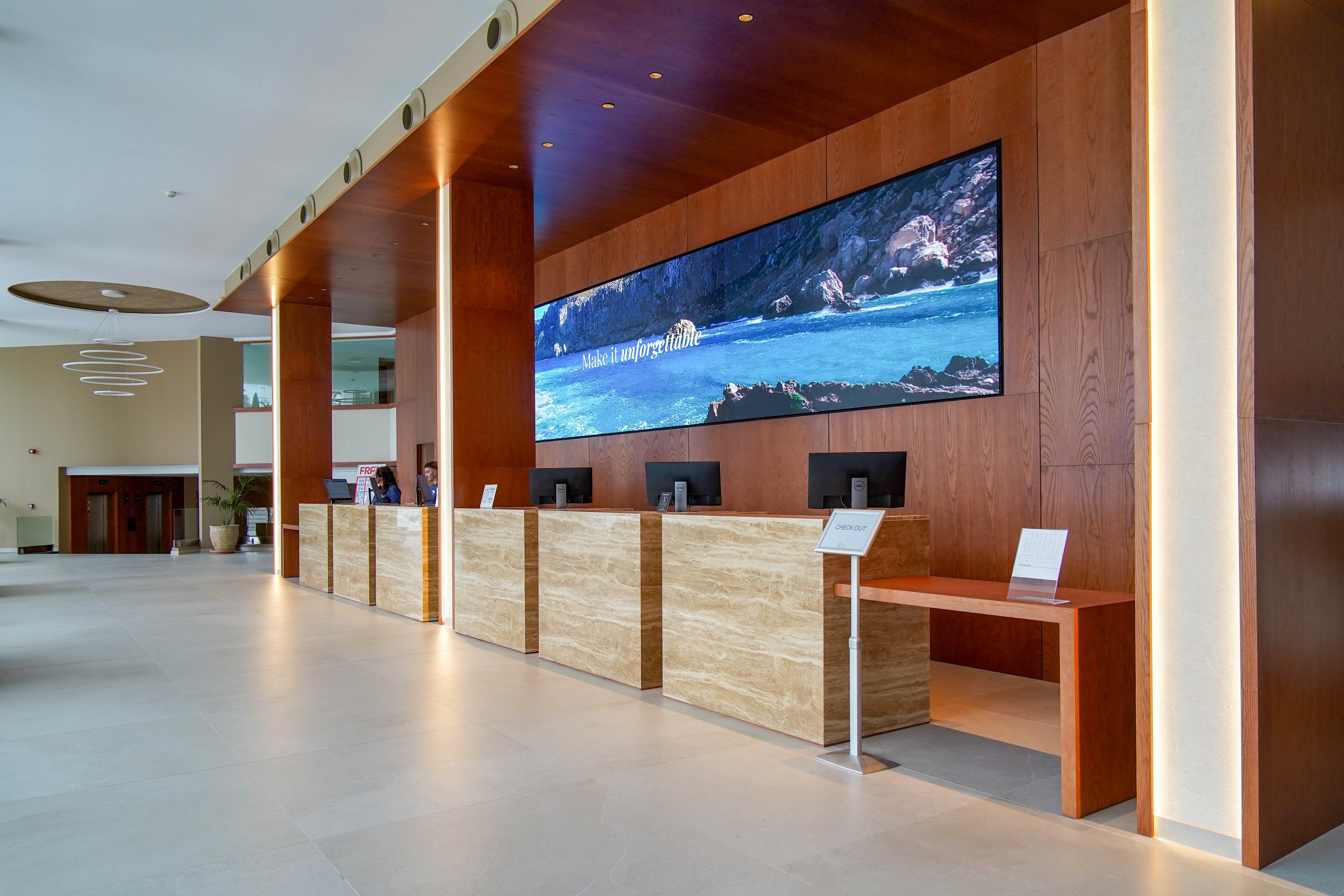 A spacious, modern hotel reception area features a long wooden and stone front desk. Multiple computer workstations are arranged along the counter, each with small information stands. A large horizontal digital screen above the desk displays a scenic coastal landscape with blue water and rocky cliffs. Warm recessed lighting and minimalist ceiling fixtures enhance the contemporary interior design. The lobby appears quiet and empty, emphasizing clean lines and open space.