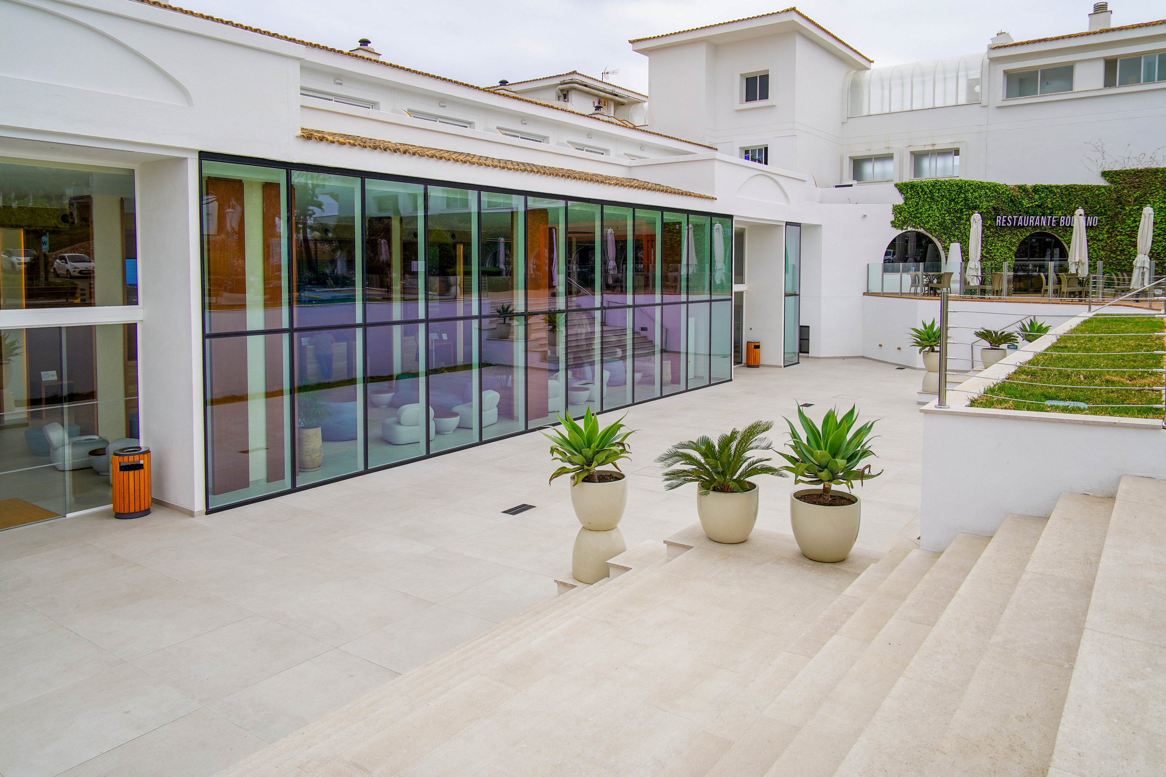 A contemporary hotel exterior features a sleek glass facade and clean white architecture. The scene shows a spacious terrace with light stone flooring and wide steps leading down to potted plants. Large windows reveal a modern interior, while a green rooftop garden adds contrast to the neutral tones. The atmosphere is calm and minimal, suggesting a resort or hospitality setting.