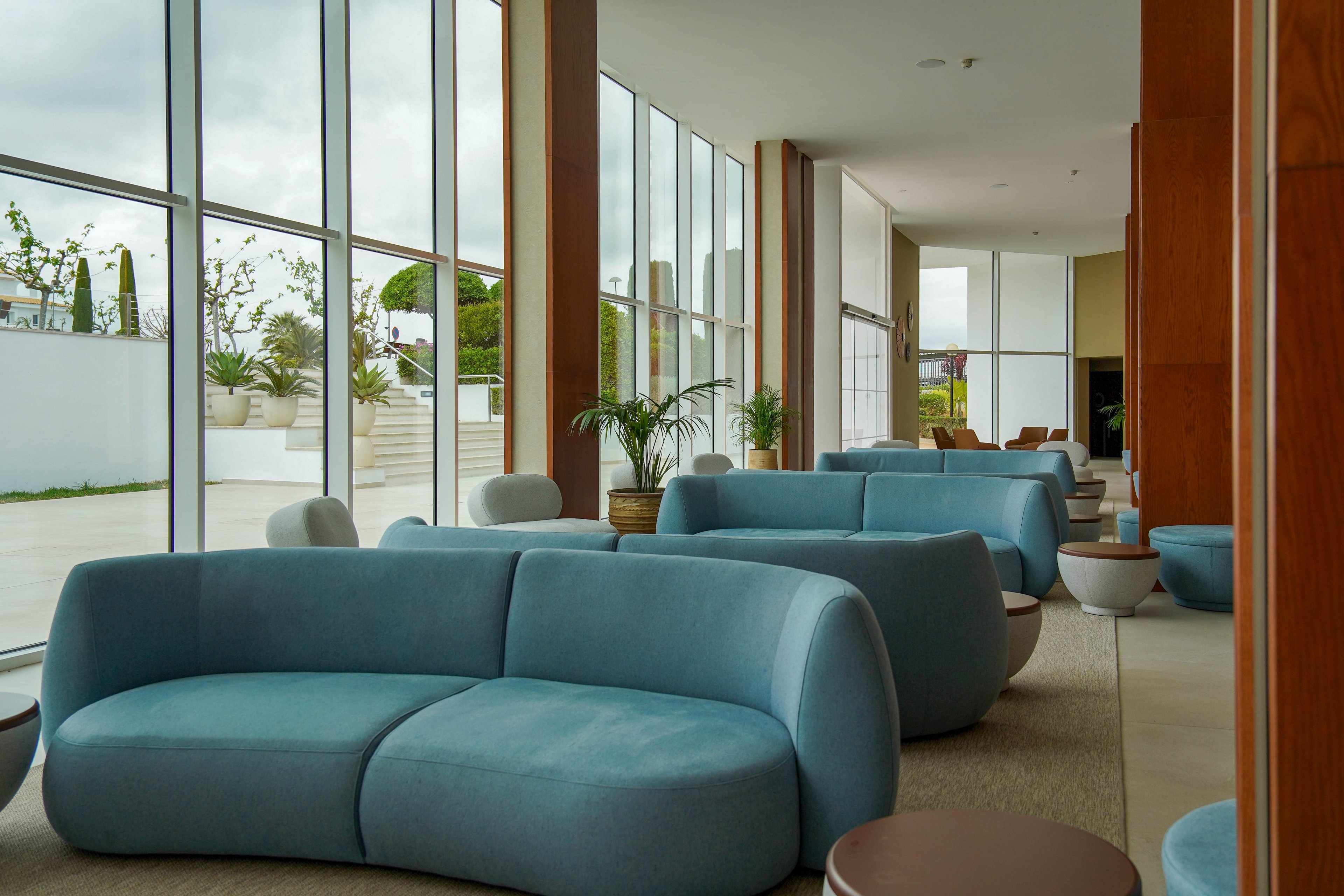 A contemporary lounge area features curved blue sofas arranged along large floor-to-ceiling windows. Natural light floods the space, highlighting minimalist furniture and neutral-toned side tables. Potted plants add greenery and soften the clean architectural lines. The overall atmosphere is calm and inviting, suitable for relaxation or informal meetings.
