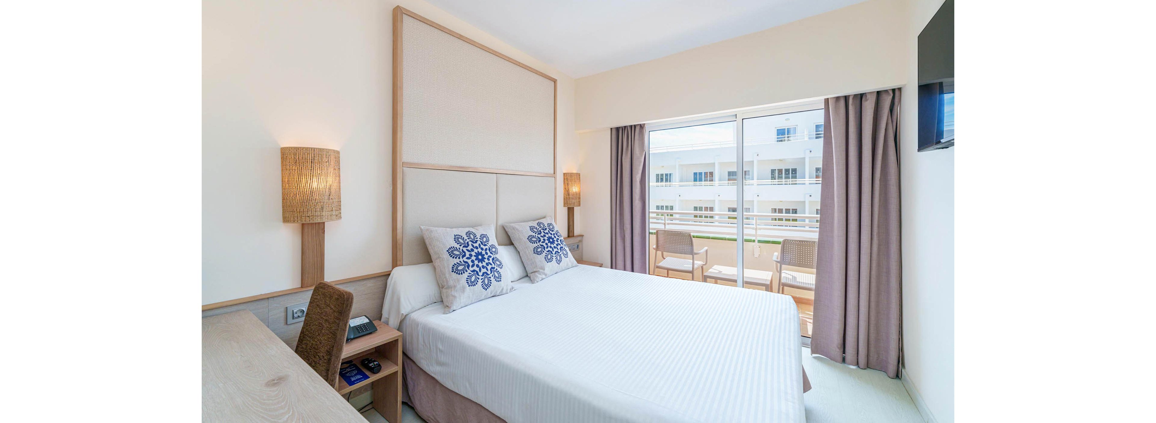 A neatly made double bed with white linens and decorative blue floral pillows is the focal point of this modern hotel room. The space features light wood furniture, including a desk and chair, and warm woven wall lamps on either side of the bed. Floor-to-ceiling glass doors open onto a balcony with outdoor chairs, revealing a view of neighboring white buildings. Soft neutral walls and curtains create a clean, airy atmosphere filled with natural light.