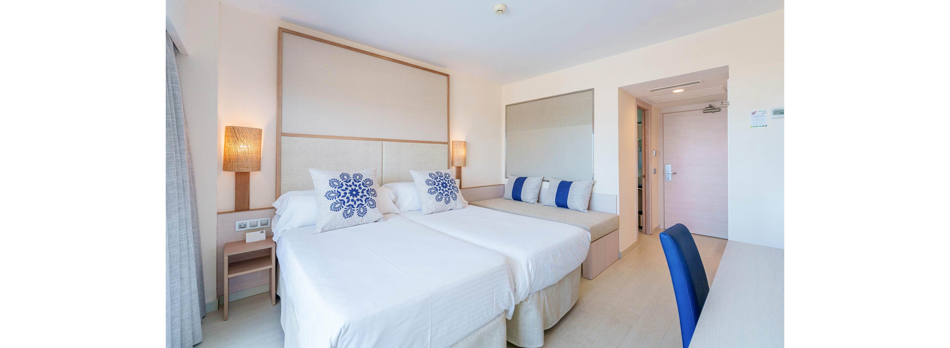 A spacious hotel room features two neatly made twin beds with white linens and decorative blue floral pillows. The setting is clean and contemporary, with a built-in sofa along the wall, accent pillows, and soft lighting from bedside lamps. The room has a neutral color palette with blue accents, a large mirror, and a visible entryway. No visible text or numeric values are present in the image.