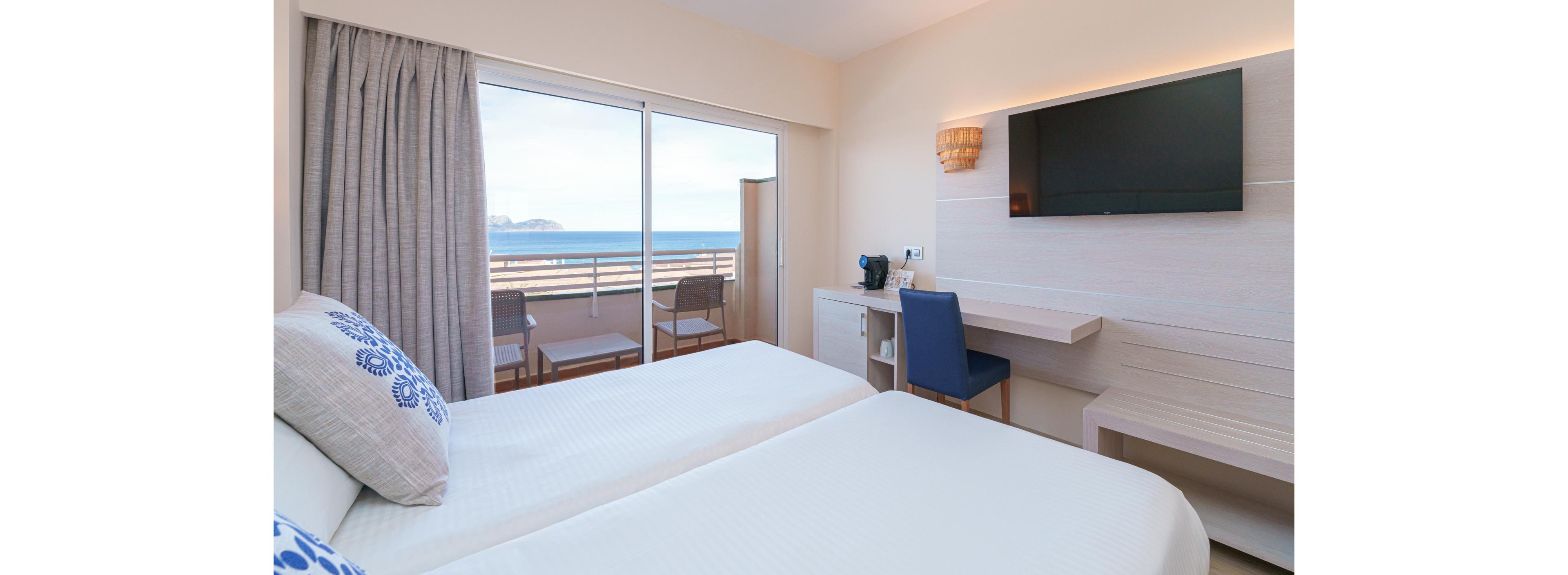 A bright, contemporary hotel room features two twin beds with white linens and blue accent pillows. The room includes a wall-mounted flat-screen TV, a small desk with a blue chair, and a coffee maker on a compact counter. Sliding glass doors open to a balcony with two chairs and a table, offering a scenic view of the ocean and distant landforms. The overall palette is light and airy, creating a relaxing atmosphere.