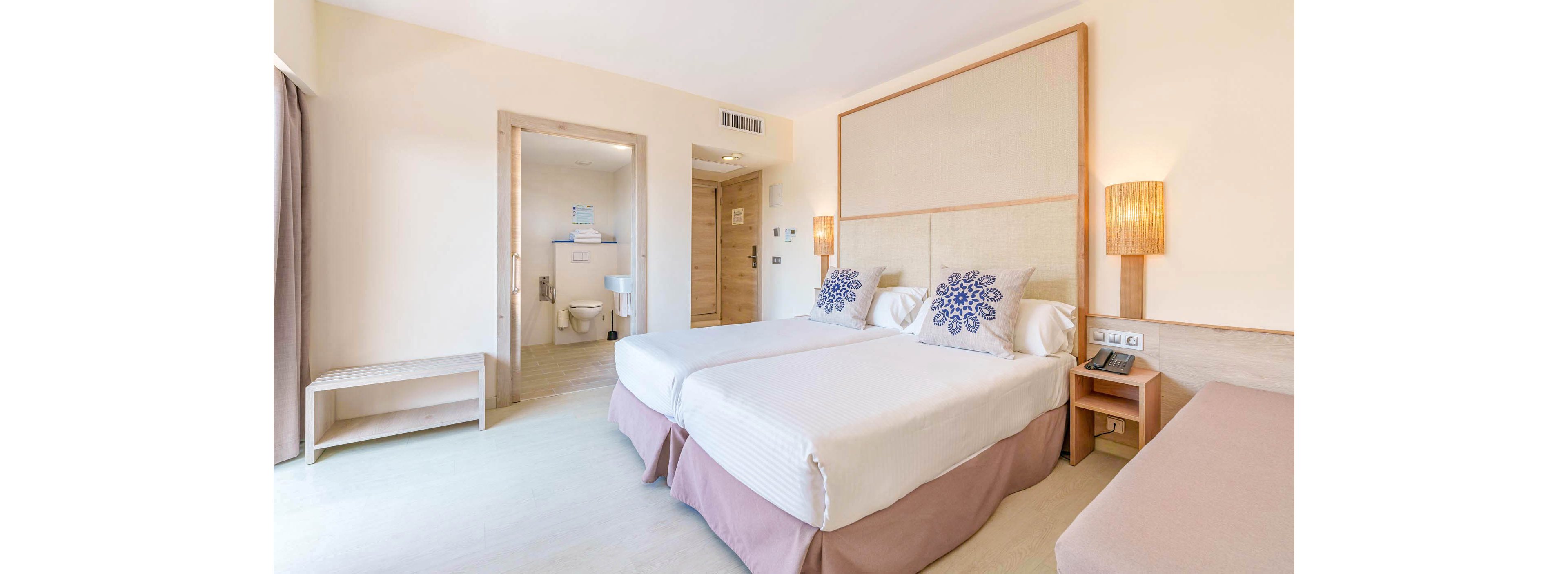 A spacious hotel room features two neatly made twin beds with decorative pillows and a light beige color palette. The room includes a bedside table, wall-mounted lamps, and an open view into a clean, well-lit bathroom. Minimalist furnishings and soft natural light create a calm and inviting atmosphere, ideal for travelers or guests seeking comfort.