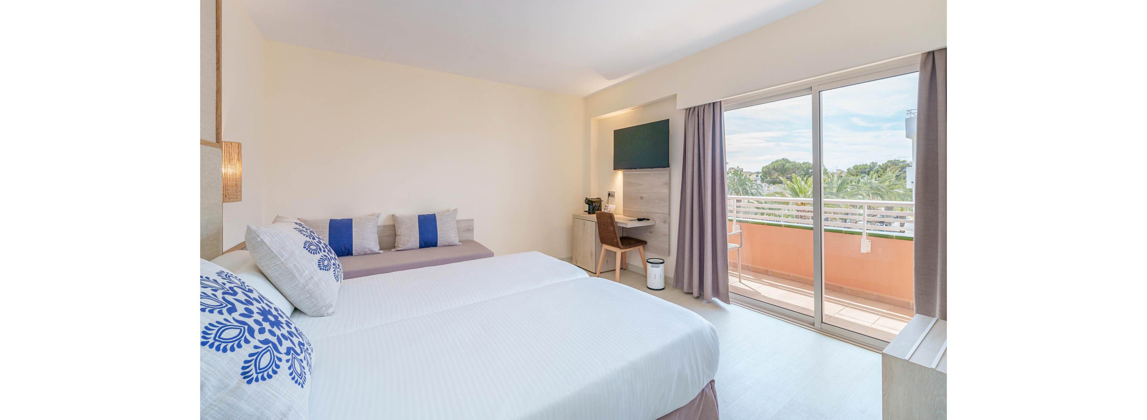 A spacious hotel room features a large bed with white linens and decorative pillows, a small sofa, and a work desk with a chair. Natural light fills the room through a sliding glass door that opens to a balcony with a view of greenery. The decor is minimalist with soft neutral tones and a touch of blue on the pillows, creating a calm and inviting atmosphere. A flat-screen TV is mounted on the wall, and a waste bin is placed near the desk. No visible text or numbers are present in the image.