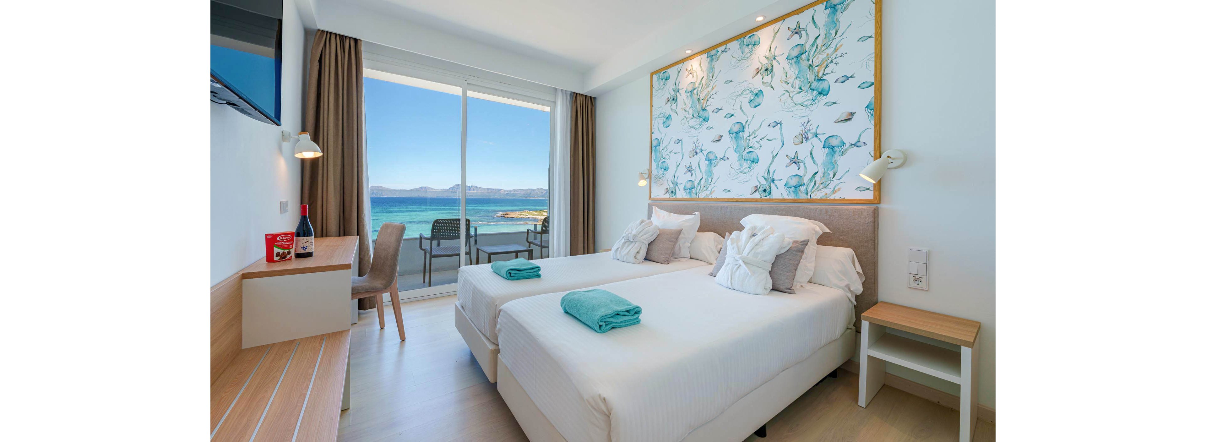 A bright modern hotel room features two neatly made single beds with folded teal towels placed on top. Large sliding glass doors open to a balcony overlooking a calm blue sea and distant mountains. Minimalist wooden furniture, including a desk, chair, and bench, complements the light flooring and neutral walls. A large decorative artwork above the beds and floor-to-ceiling curtains add a stylish touch to the serene coastal setting.