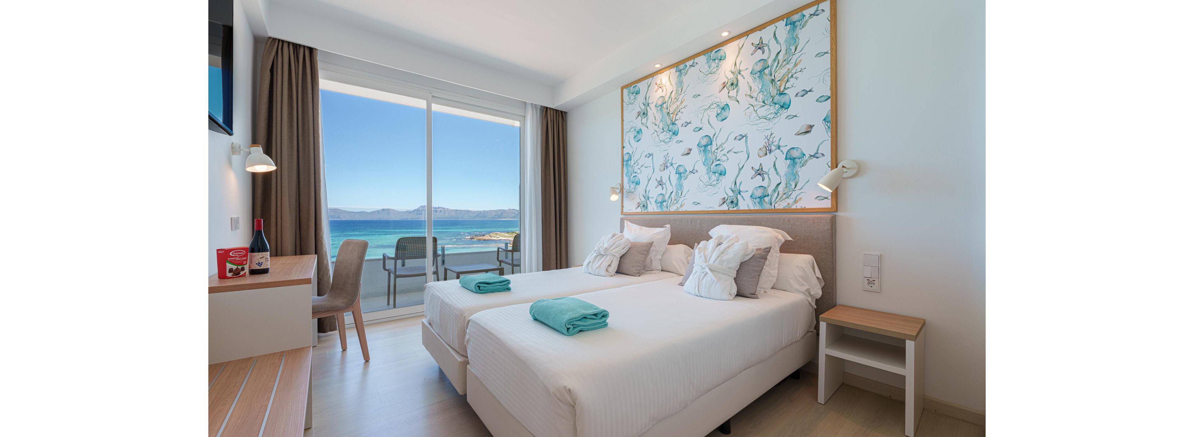 Modern hotel room features two neatly made twin beds with white linens and folded turquoise towels. A large sliding glass door opens to a balcony overlooking a vivid blue sea and distant mountains. Soft neutral tones, light wood furniture, and an abstract ocean-themed wall art create a fresh, coastal atmosphere. A desk area with a chair and a bottle of wine adds a touch of comfort and relaxation.