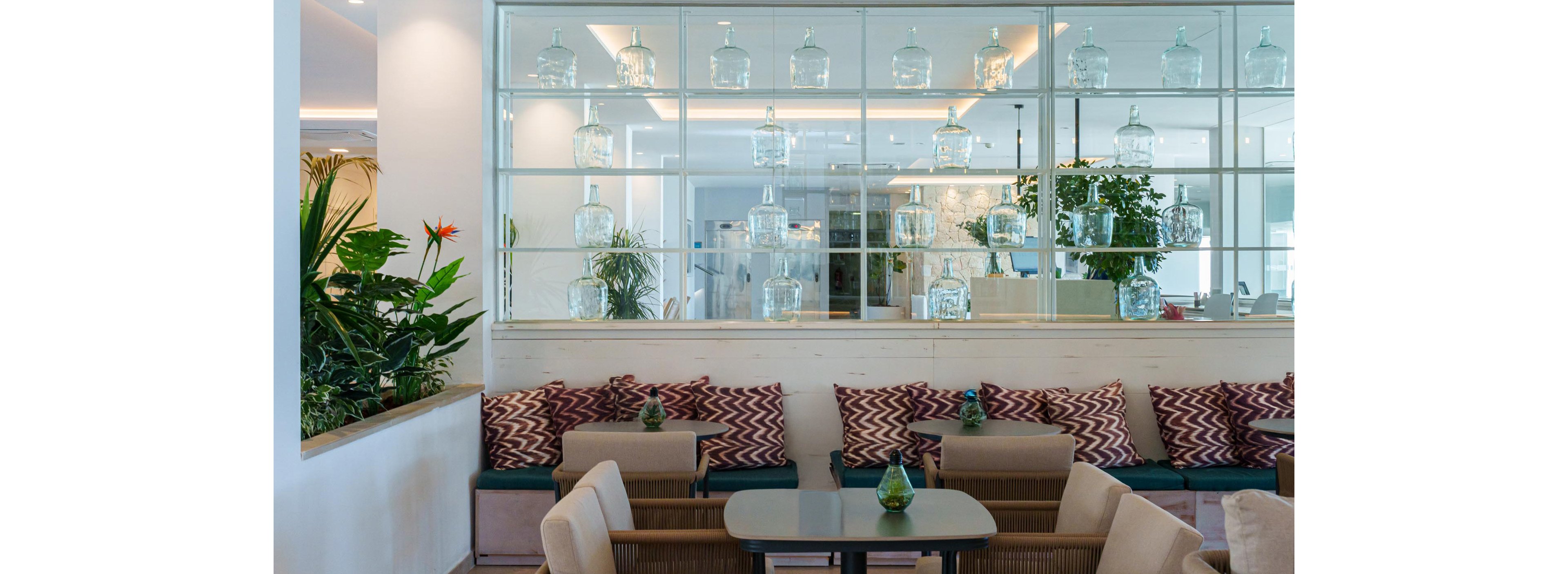 A contemporary lounge features cushioned bench seating with patterned pillows and small tables. The setting is bright and airy, with a glass partition displaying multiple clear jars and lush green plants adding freshness. The color palette includes soft neutrals, greens, and pops of red from decorative pillows, creating a relaxed and inviting atmosphere.