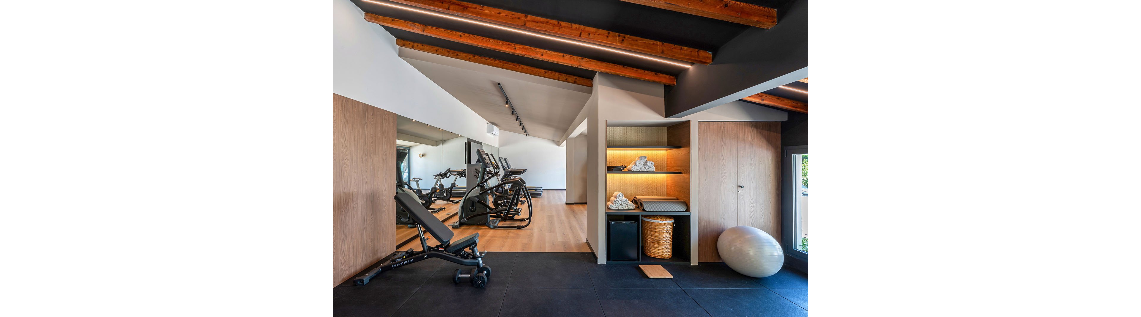 Contemporary home gym interior featuring cardio machines and weight equipment. The space includes treadmills, an exercise bike, and a weight bench arranged on a wooden floor. A large wall mirror visually expands the room, while open shelving with towels and storage baskets adds warmth and functionality. Exposed wooden ceiling beams and soft lighting create a clean, inviting workout environment.