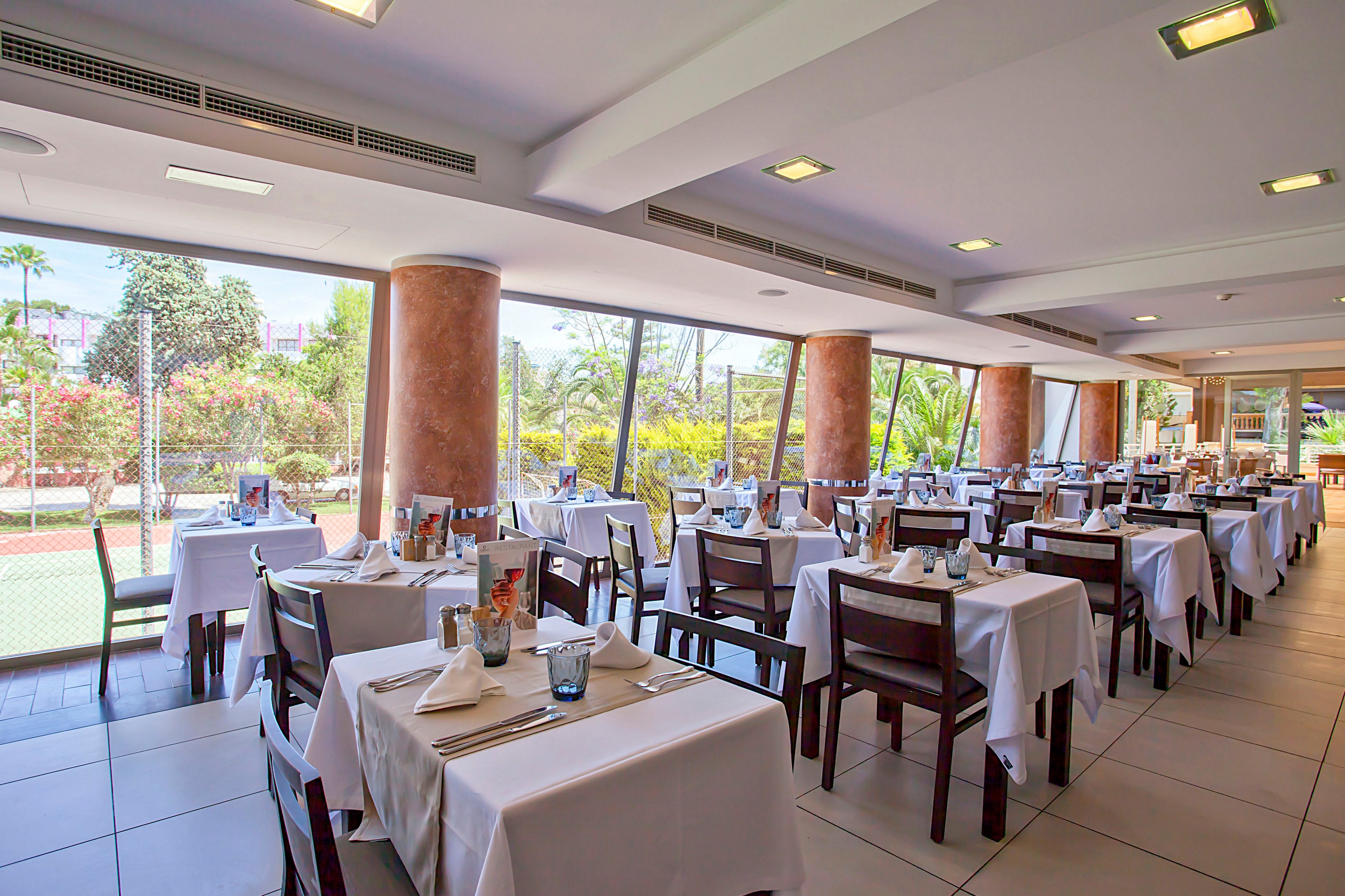 A modern restaurant dining room is set with neatly arranged tables covered in white tablecloths. Wooden chairs surround the tables, each prepared with folded napkins, glassware, and simple place settings. Large floor-to-ceiling windows line one side of the room, revealing a lush outdoor garden and tennis court. The space is brightly lit with recessed ceiling lights, creating an airy and welcoming atmosphere.