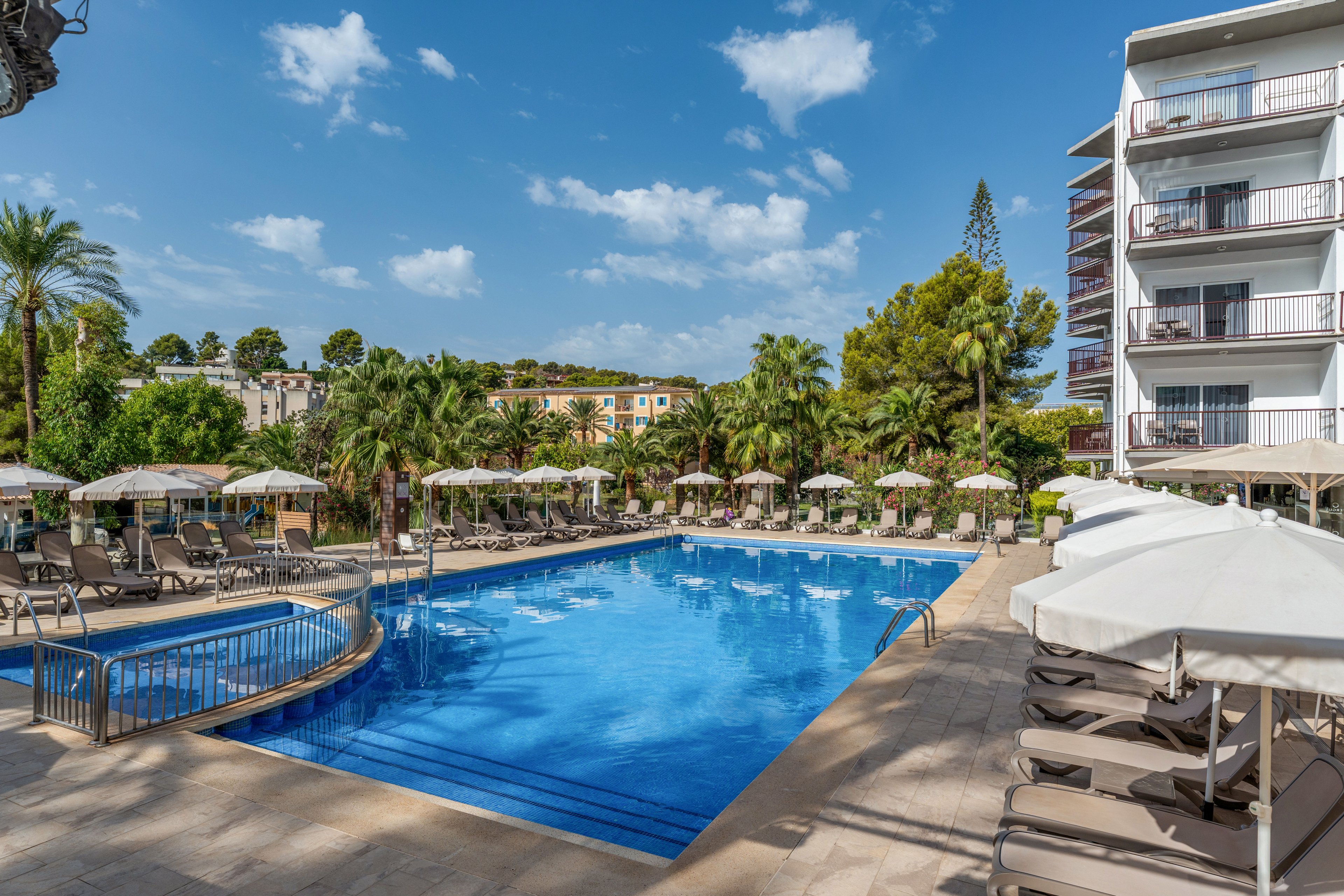 A clear blue swimming pool sits at the center of a hotel courtyard under a bright sky. Numerous sun loungers and beige umbrellas line the poolside, creating a relaxed resort atmosphere. A multi-story hotel building with balconies stands to the right, while lush green palm trees and other vegetation surround the area. The scene suggests a tranquil vacation setting on a warm day.