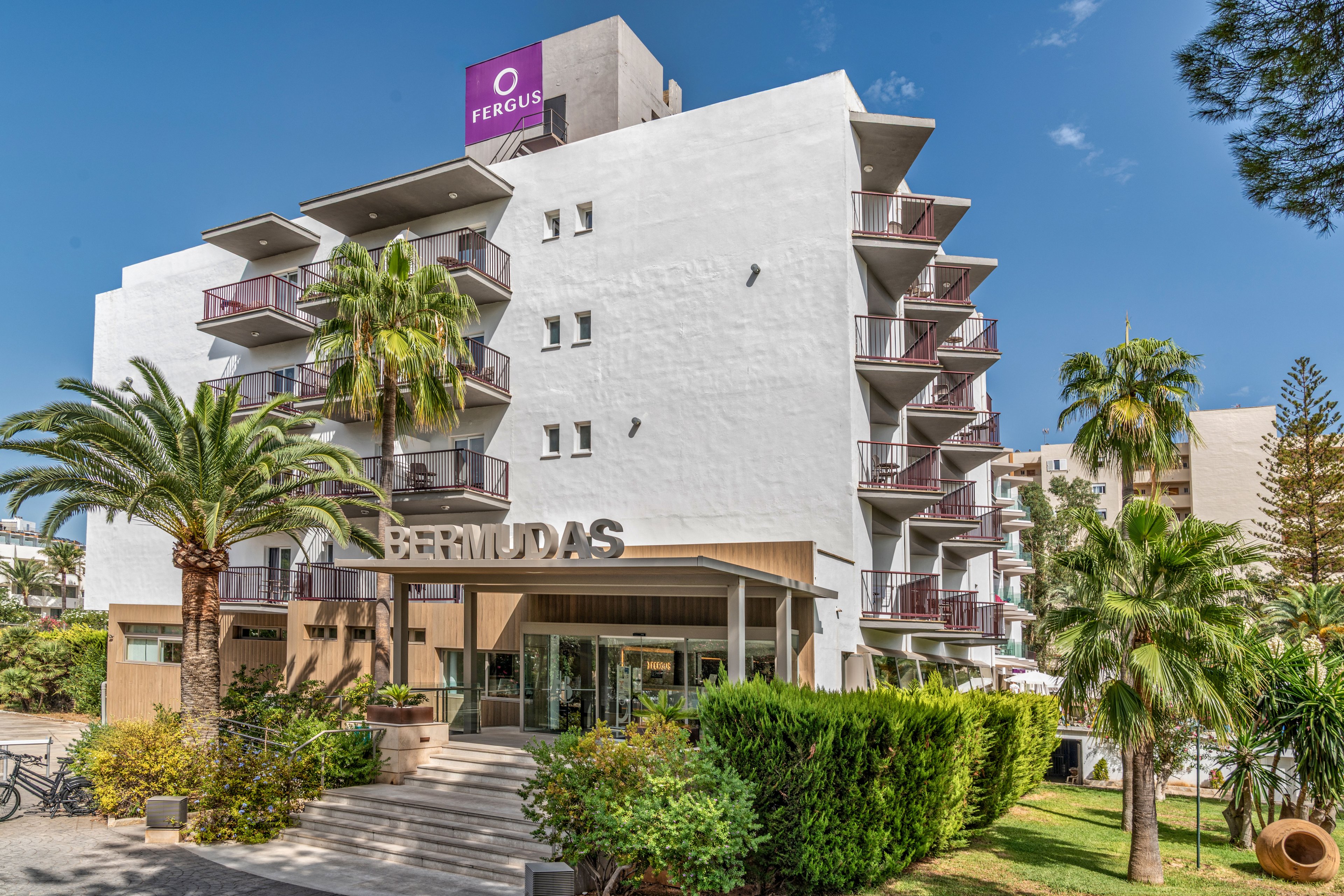 Contemporary multi-story hotel building named Bermudas with a prominent entrance canopy. The facade features multiple balconies with railings and a clean white exterior. Lush palm trees, shrubs, and landscaped greenery surround the front of the property under a bright blue sky. The scene suggests a sunny vacation destination in a warm, coastal or resort area.