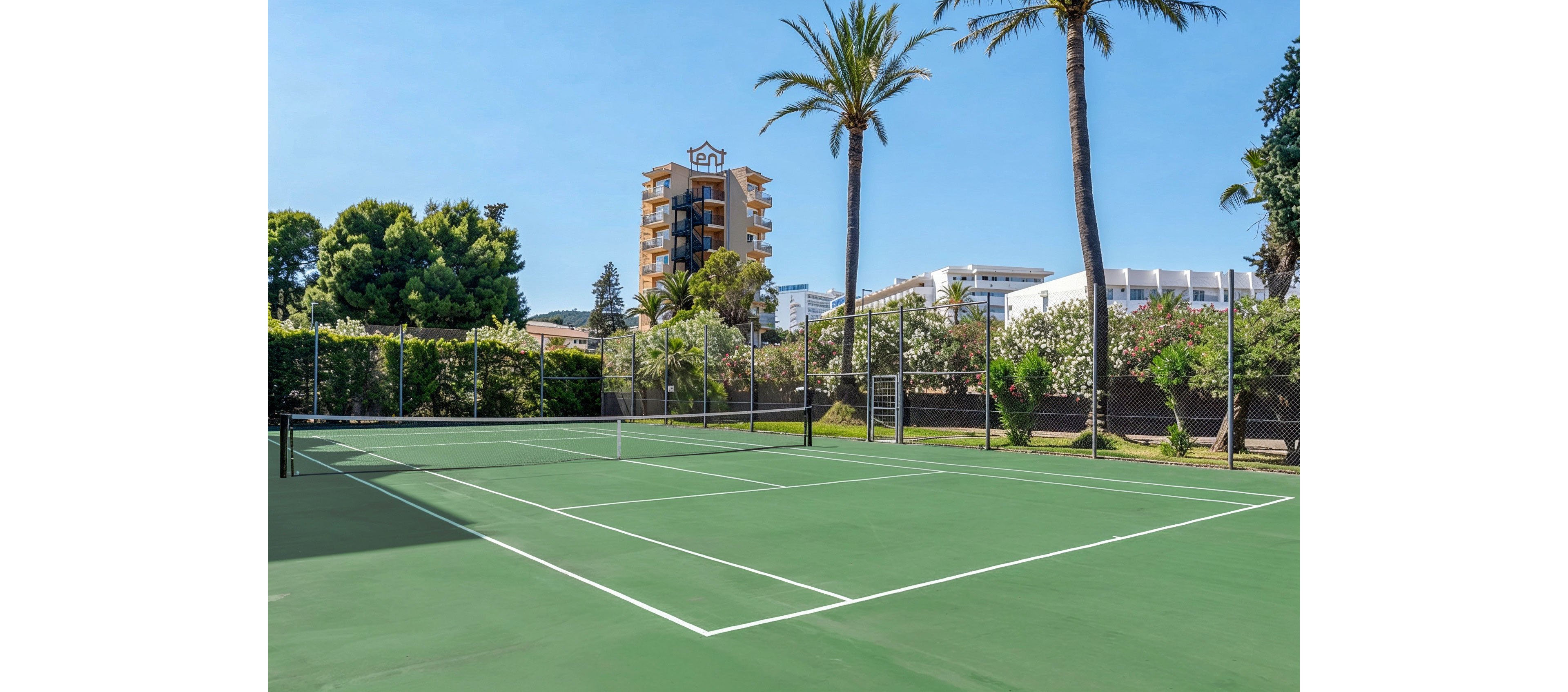 An empty green tennis court is shown under a clear blue sky. The court is surrounded by a tall chain-link fence and lush greenery, including palm trees and hedges. In the background, mid-rise apartment or hotel buildings are visible, suggesting a resort or residential area. The bright daylight and clean court lines create a fresh, inviting atmosphere for outdoor sports.