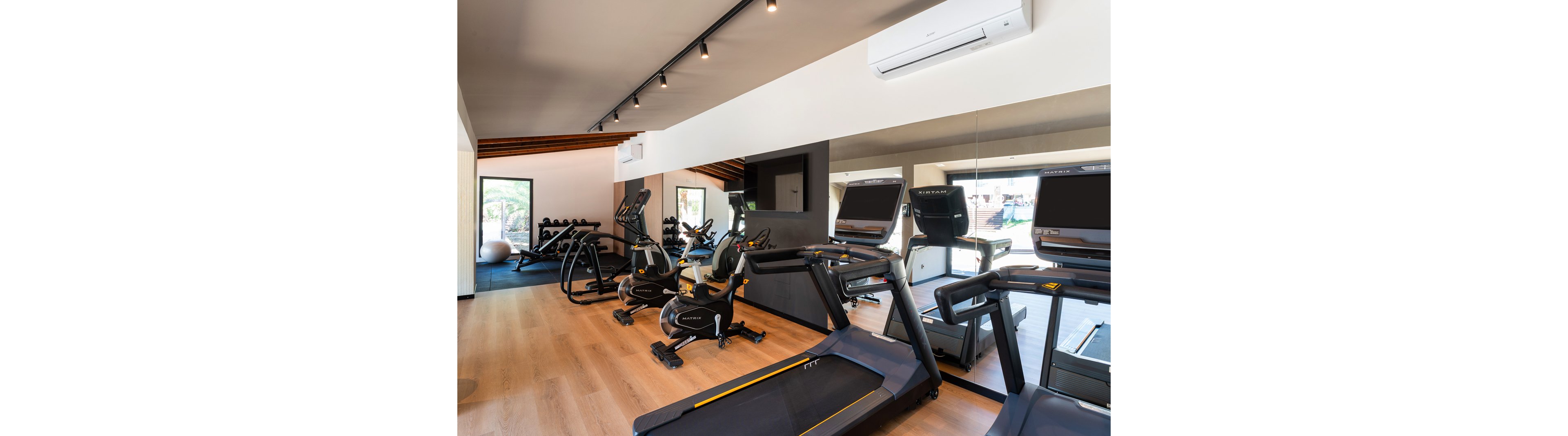 A bright, modern gym room features several treadmills and stationary bikes neatly arranged on a wooden floor. Large mirrors along one wall reflect the exercise machines and expand the sense of space. Natural light enters through a window, highlighting the clean, minimalist interior and neutral color palette. The room appears quiet and ready for individual workouts or cardio training sessions.