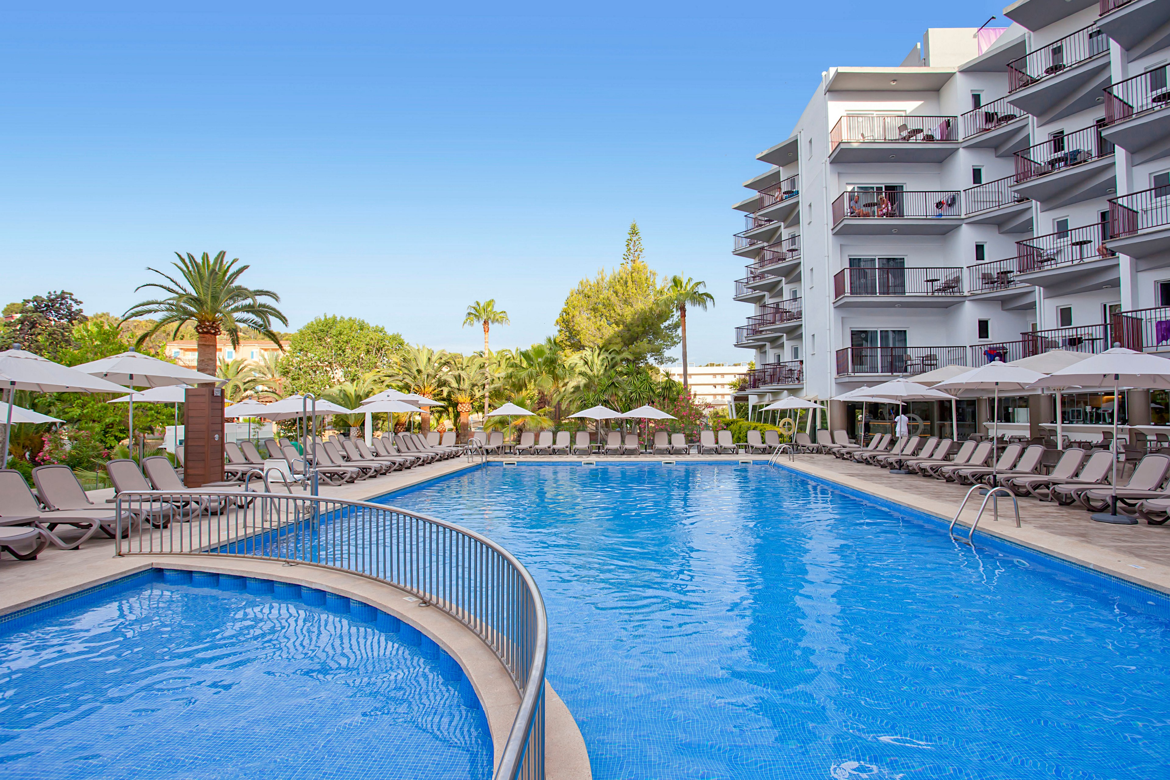 Large outdoor swimming pool at a modern resort hotel is surrounded by numerous sun loungers and white umbrellas. A multi-story building with balconies overlooks the pool area on the right side. Lush green palm trees and landscaped gardens frame the scene under a clear blue sky. The calm water and empty chairs suggest a peaceful, relaxing vacation setting.