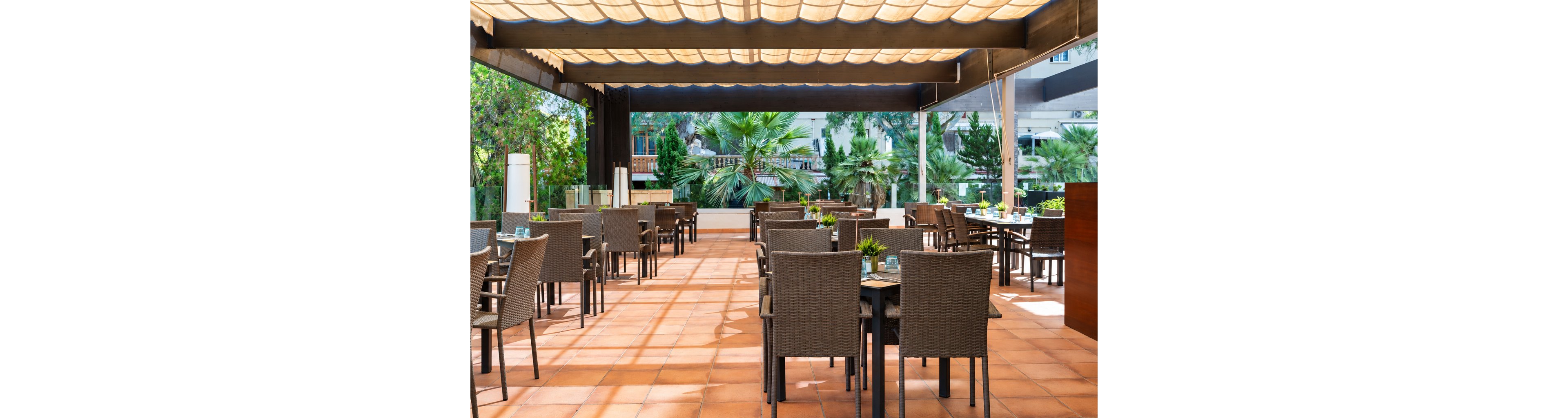 An open-air restaurant terrace features neatly arranged tables and wicker chairs on a terracotta-tiled floor. A large pergola with a light-colored canopy casts soft shadows across the dining area. Green plants and palm trees surround the space, creating a lush garden atmosphere. The setting appears calm and inviting, suitable for relaxed outdoor dining.