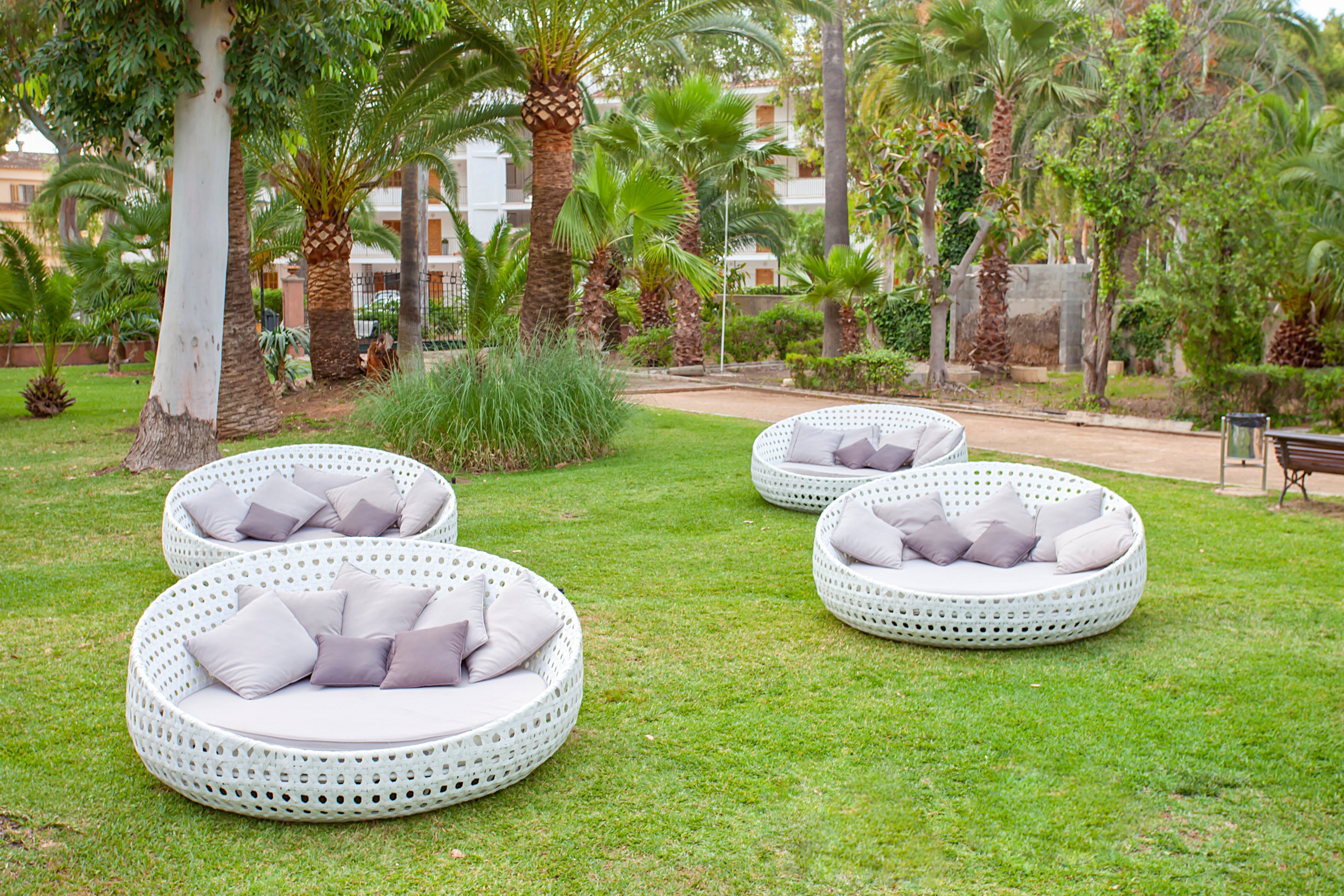 Four round white wicker lounge chairs with cushions are arranged on a manicured lawn. The seating area is set in a lush tropical garden with palm trees and dense greenery. Neutral-toned pillows in gray and beige add a soft, relaxing touch to the modern outdoor furniture. A pathway and buildings are visible in the background, suggesting a resort or landscaped public space.