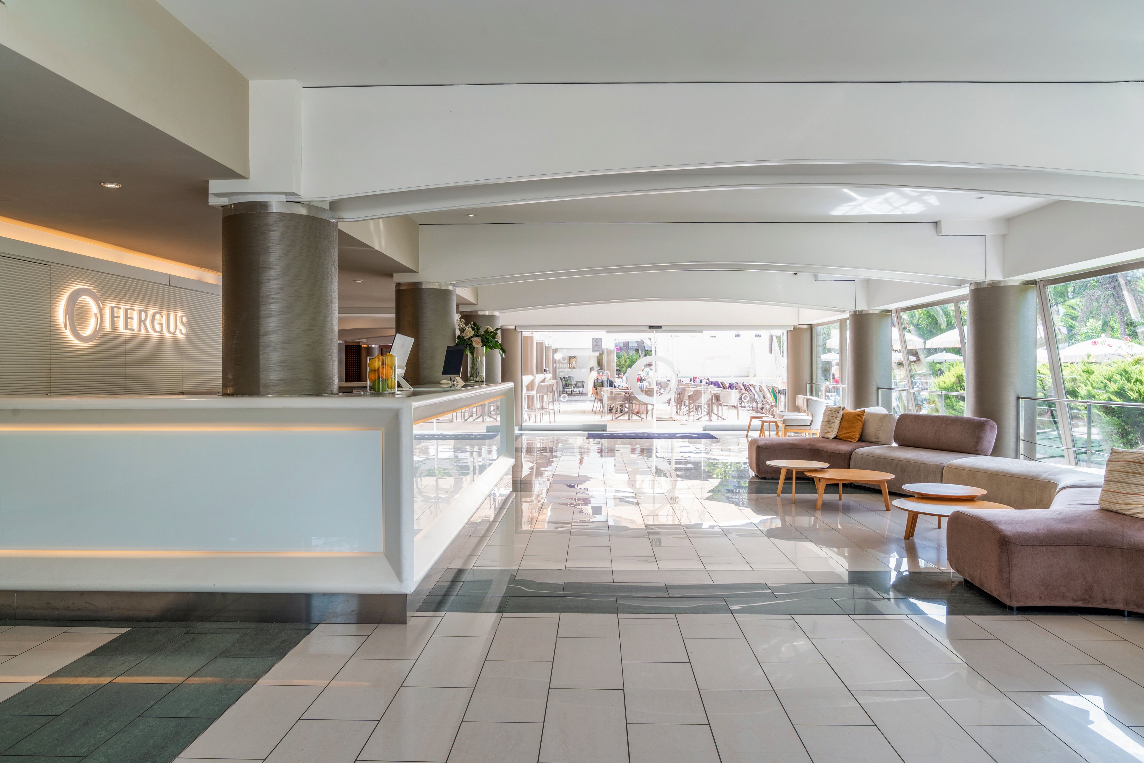 A spacious, modern hotel lobby features a sleek white reception desk on the left side. The area is filled with natural light from large windows at the far end, revealing an outdoor terrace with tables and chairs. Comfortable sofas and small round tables create a relaxed seating area along the right side. Neutral tones, clean lines, and polished tile flooring contribute to a bright, contemporary atmosphere.