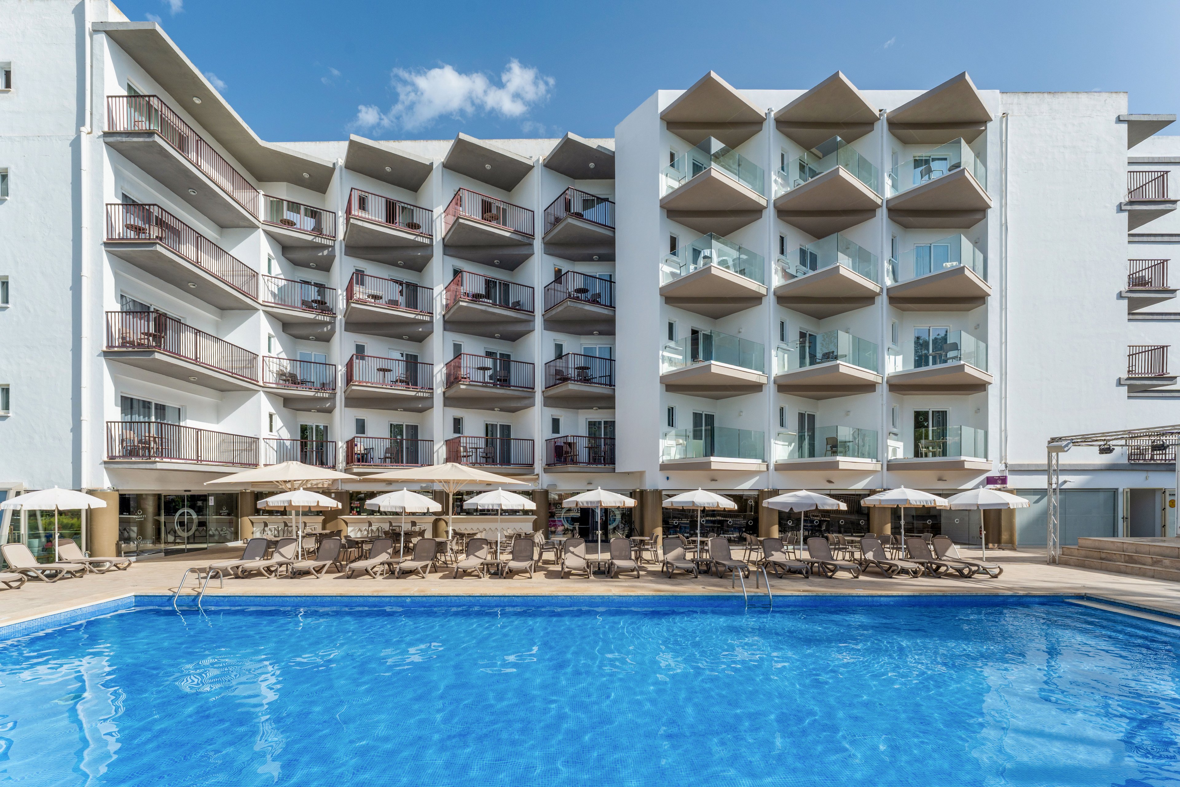A contemporary multi-story hotel building features rows of private balconies facing a large outdoor swimming pool. Sun loungers and parasols line the poolside terrace, suggesting a relaxing vacation setting. The clear blue water reflects the bright sky and white facade of the hotel. The scene conveys a sunny, tranquil resort atmosphere ideal for leisure travel.