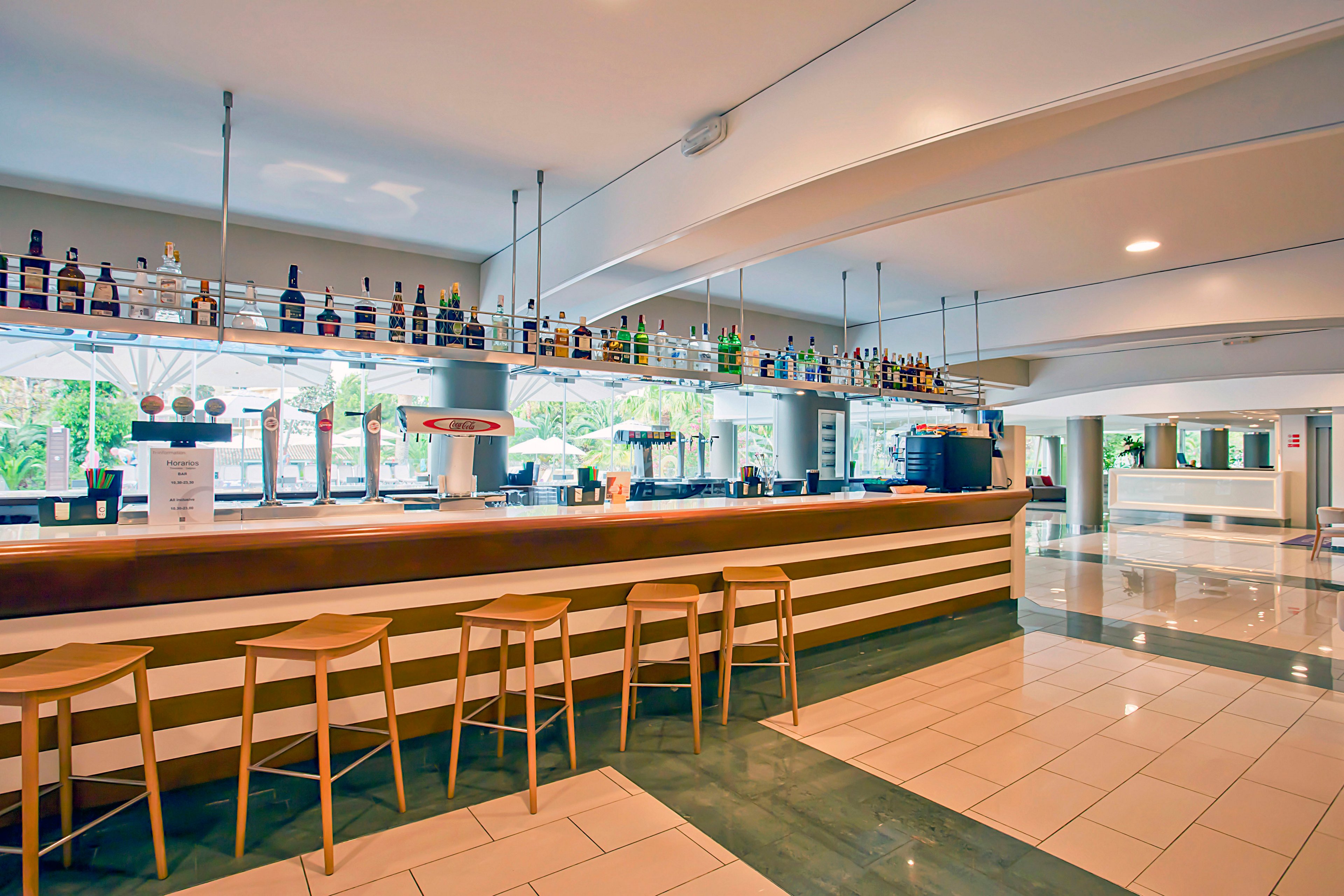 A contemporary bar interior features a long wooden counter with several minimalist stools neatly arranged in front. Bottles of liquor are displayed on glass shelves suspended above the bar, with large windows revealing greenery outside. The space is bright and airy, with polished tile floors and clean architectural lines. No people are present, emphasizing the calm, orderly atmosphere of the venue.