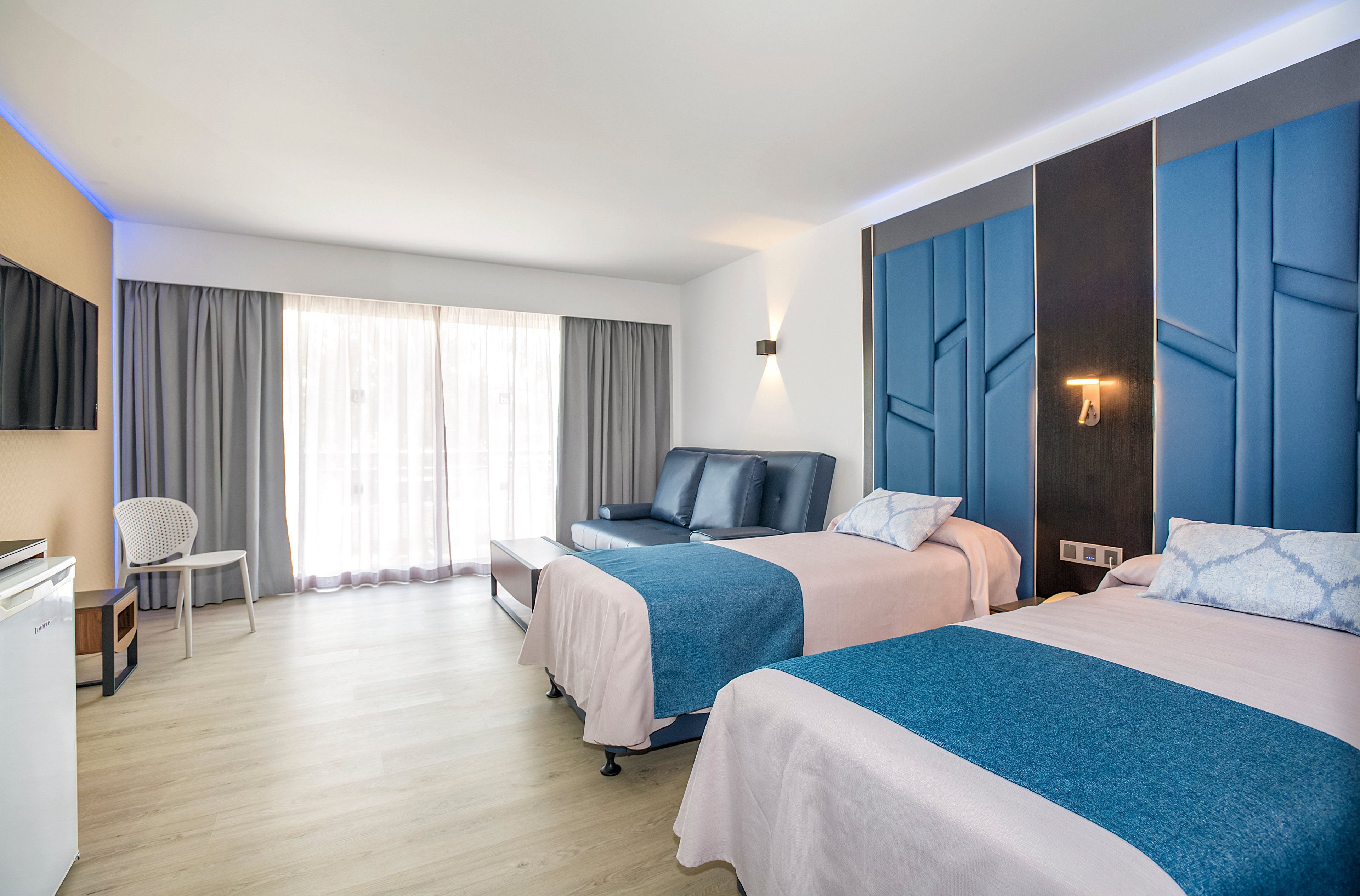 A contemporary hotel room featuring two twin beds with blue and white bedding. The setting includes a seating area, wall-mounted TV, and large windows with sheer curtains allowing natural light. The room has a clean and minimalist design with wooden flooring and soft lighting.