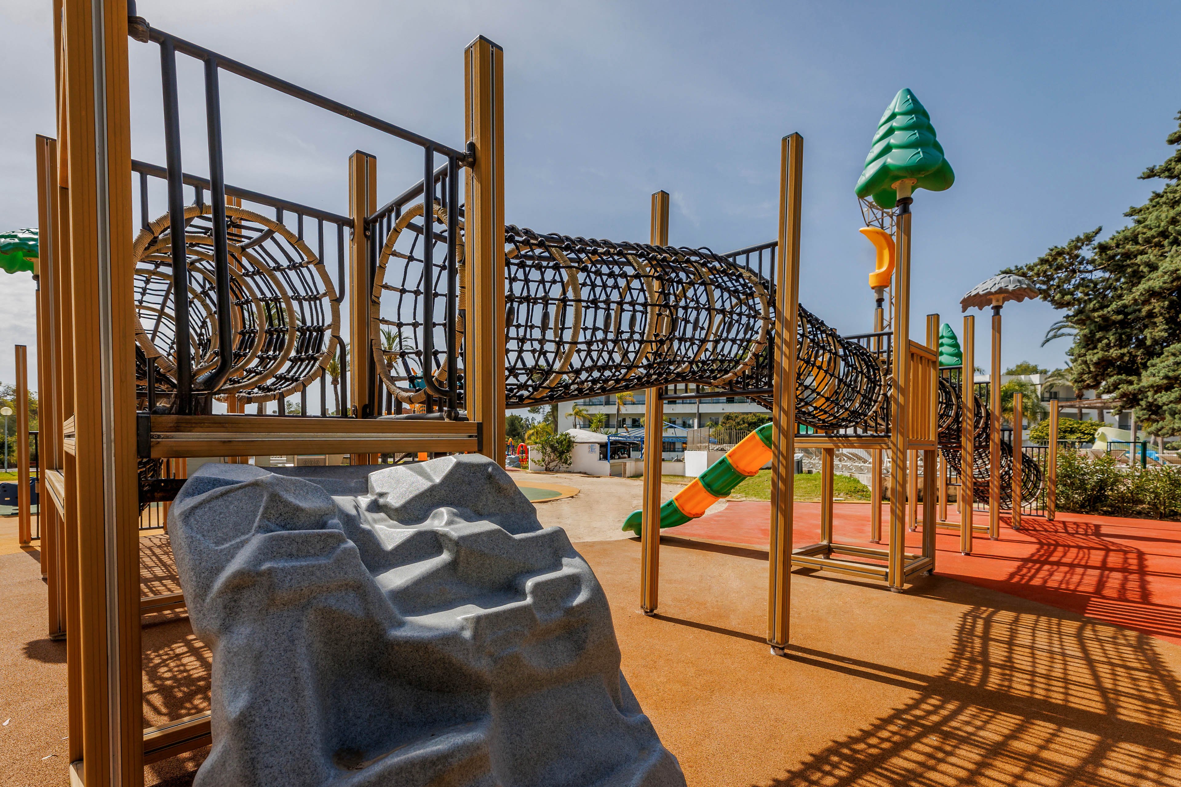 An outdoor playground features a large tubular climbing tunnel supported by tall posts. In the foreground, a textured gray climbing rock invites children to scale its surface. Bright orange and green play structures, including slides and decorative tree elements, add color to the scene. The area is set on a soft, rubberized surface under a clear, sunny sky, suggesting a safe and inviting recreational space.