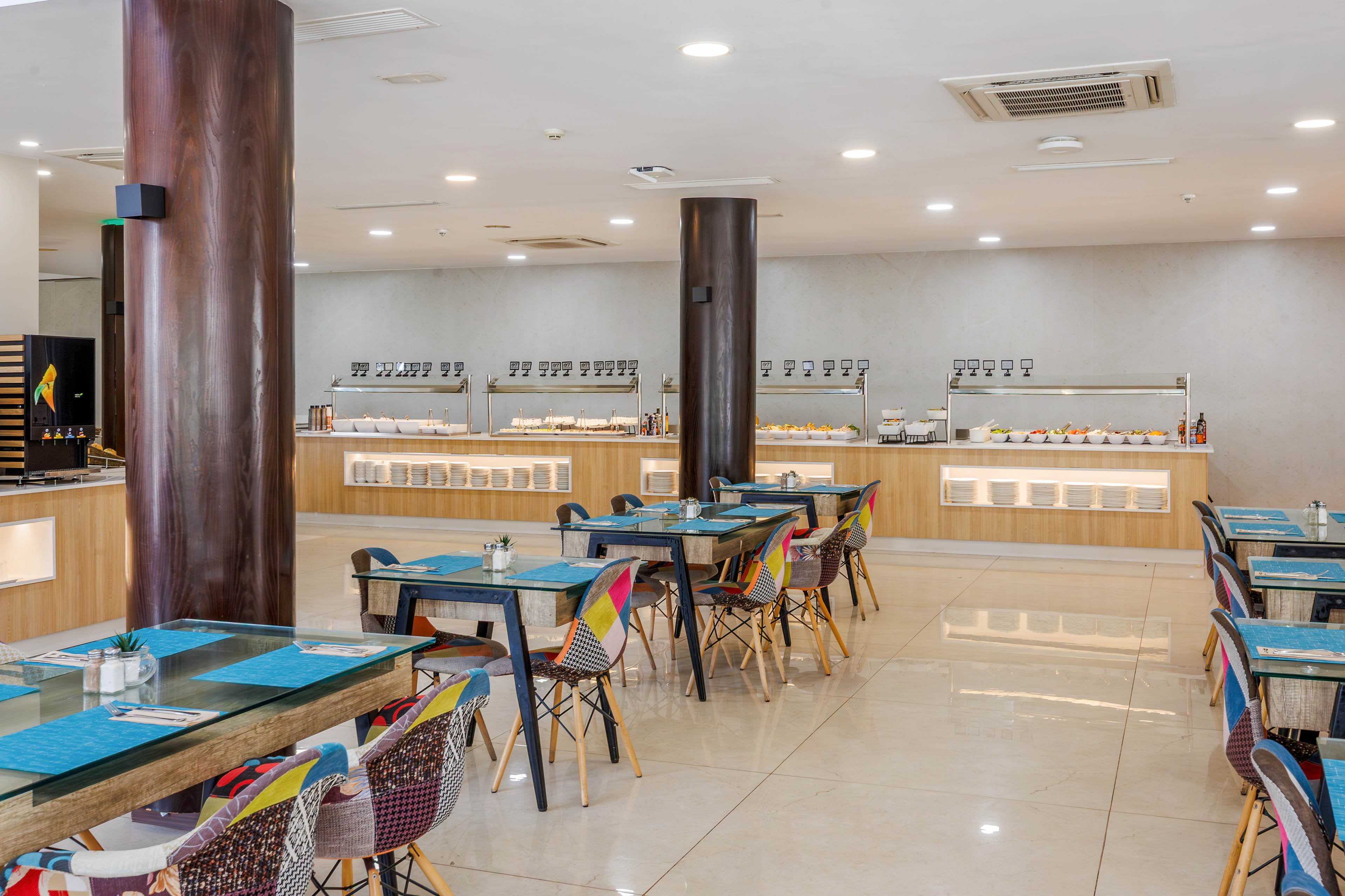 A contemporary self-service restaurant features rows of empty tables with colorful, patterned chairs and turquoise placemats. In the background, a long buffet counter displays covered dishes and food trays under warm lighting. The space is bright and clean, with glossy tiled floors, wooden columns, and a neutral wall. Ceiling lights and air conditioning units contribute to the modern, comfortable dining atmosphere.
