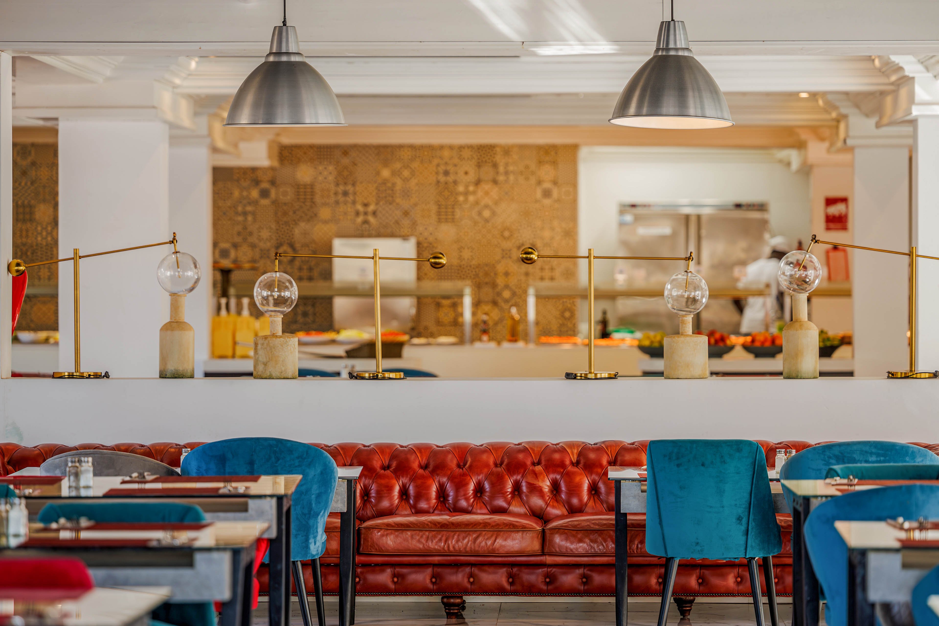 A contemporary restaurant interior features a rich brown leather tufted sofa as the central focal point. Blue upholstered chairs and neatly set tables with placemats and glassware create an inviting dining area. In the background, an open kitchen with patterned tile walls and metallic pendant lights adds visual interest. Warm lighting and decorative glass globes on brass fixtures contribute to a cozy yet modern atmosphere.