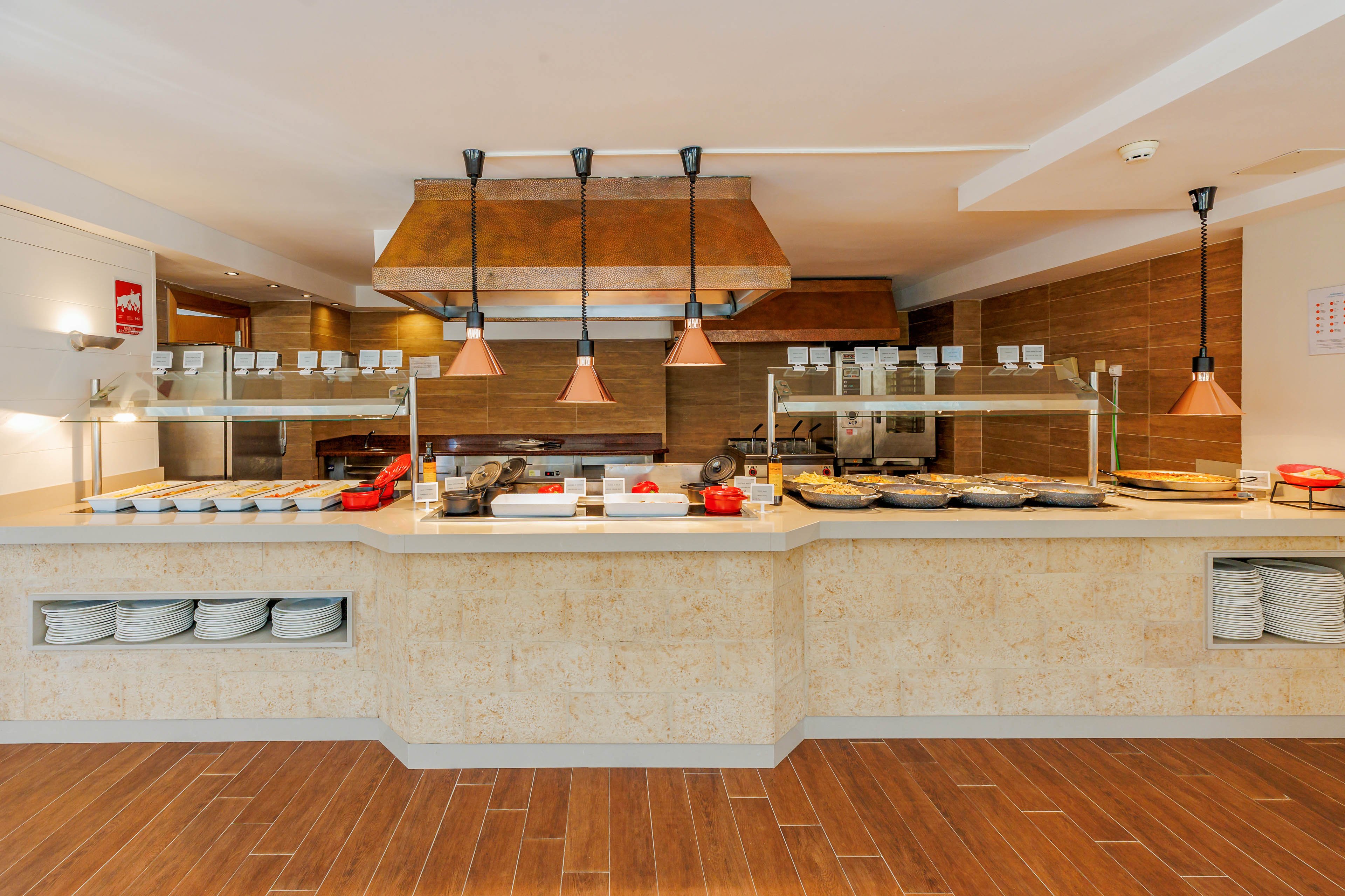 A clean, empty buffet counter is set up in a restaurant or hotel dining area. Warm pendant lights hang above the serving line, illuminating trays, chafing dishes, and glass sneeze guards. Stacks of white plates are neatly arranged in open shelves beneath the counter. The space features wood-look flooring and wood-paneled walls, creating a modern, inviting atmosphere.