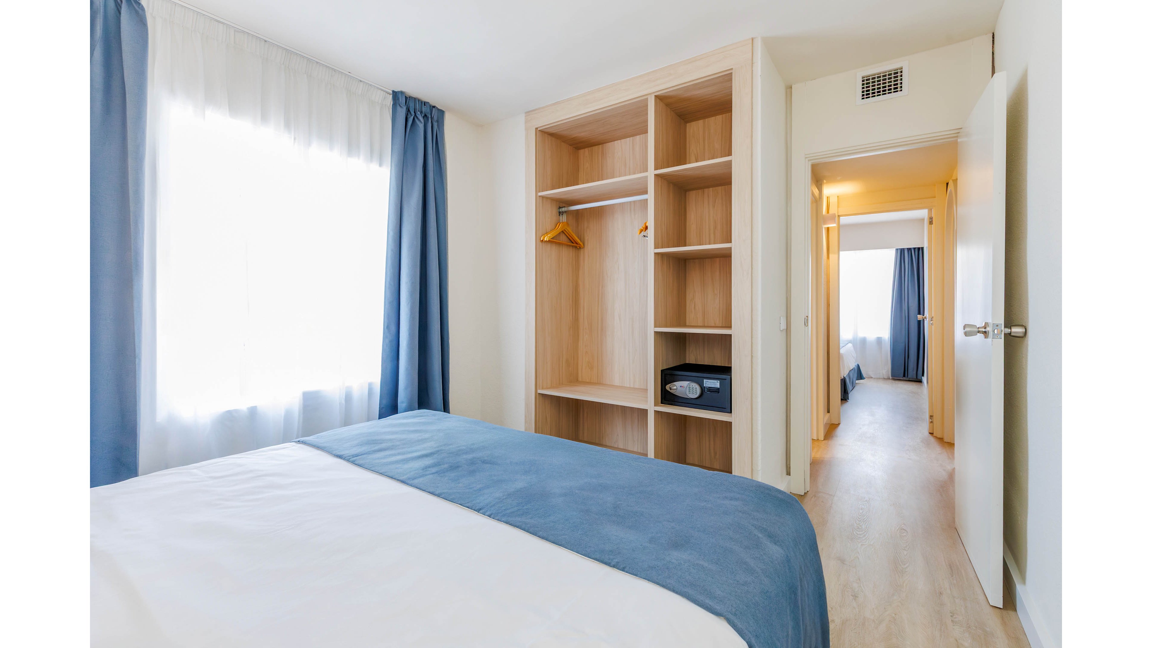 A clean, modern bedroom features a neatly made bed with white linens and a blue bed runner. An open wooden wardrobe with shelves, a hanging rail, and a small safe is positioned against the wall. Natural light streams through a large window with sheer curtains and blue drapes, creating a bright and airy atmosphere. A hallway leads to another similarly styled room, emphasizing a contemporary hotel or apartment interior.