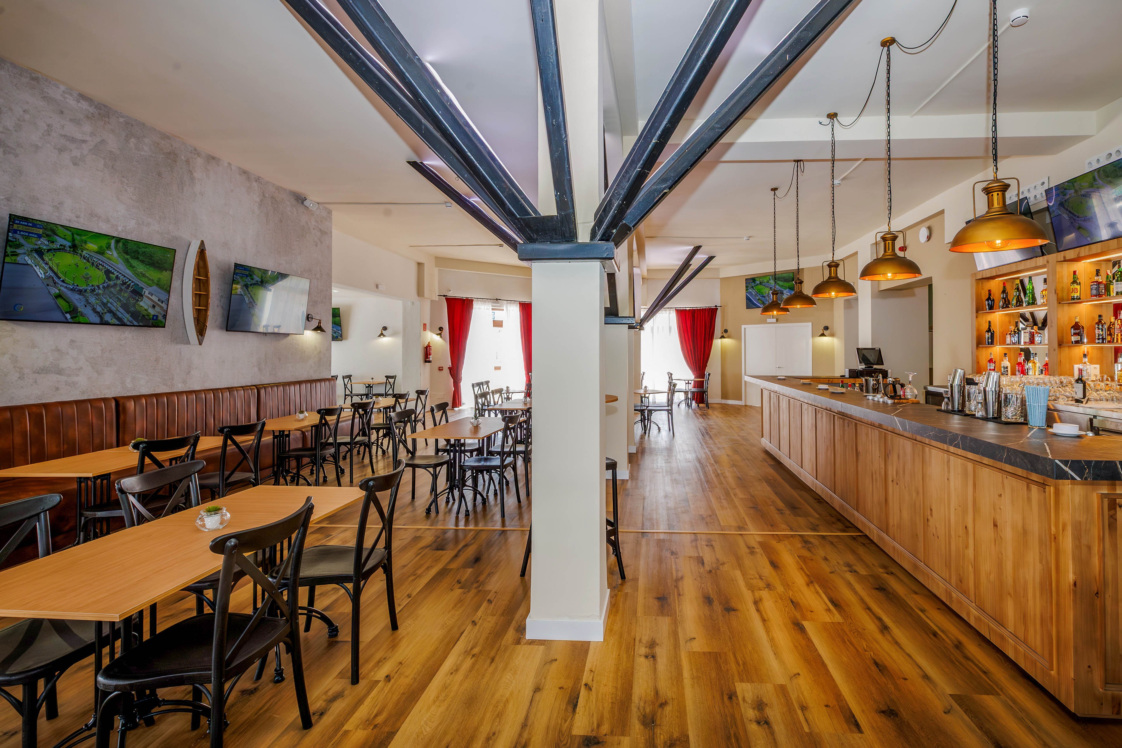 Spacious bar and restaurant interior with wooden floors and a long wood-paneled counter. Multiple tables and chairs are neatly arranged in an open dining area, with no people present. Warm pendant lights hang above the bar, illuminating bottles and glassware on the shelves. Wall-mounted screens and red curtains add visual interest to the contemporary, welcoming space.