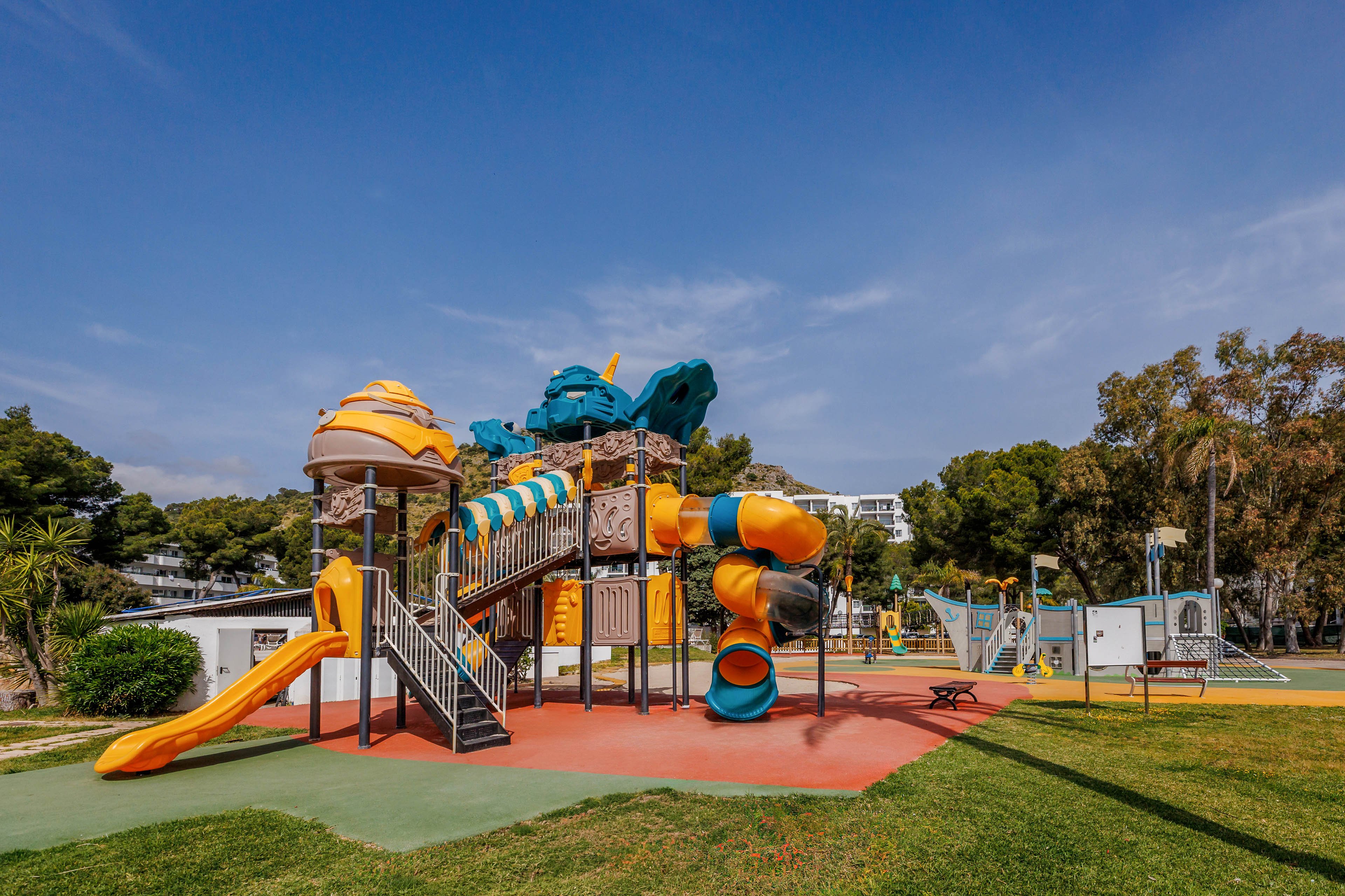 A bright, multi-level playground structure with several slides stands in an open outdoor park. The equipment features orange, yellow, and blue elements, including tunnels, stairways, and climbing sections. Surrounding the play area are green lawns, trees, and nearby park facilities under a clear blue sky. The scene suggests a family-friendly recreational space designed for children’s play and activity.