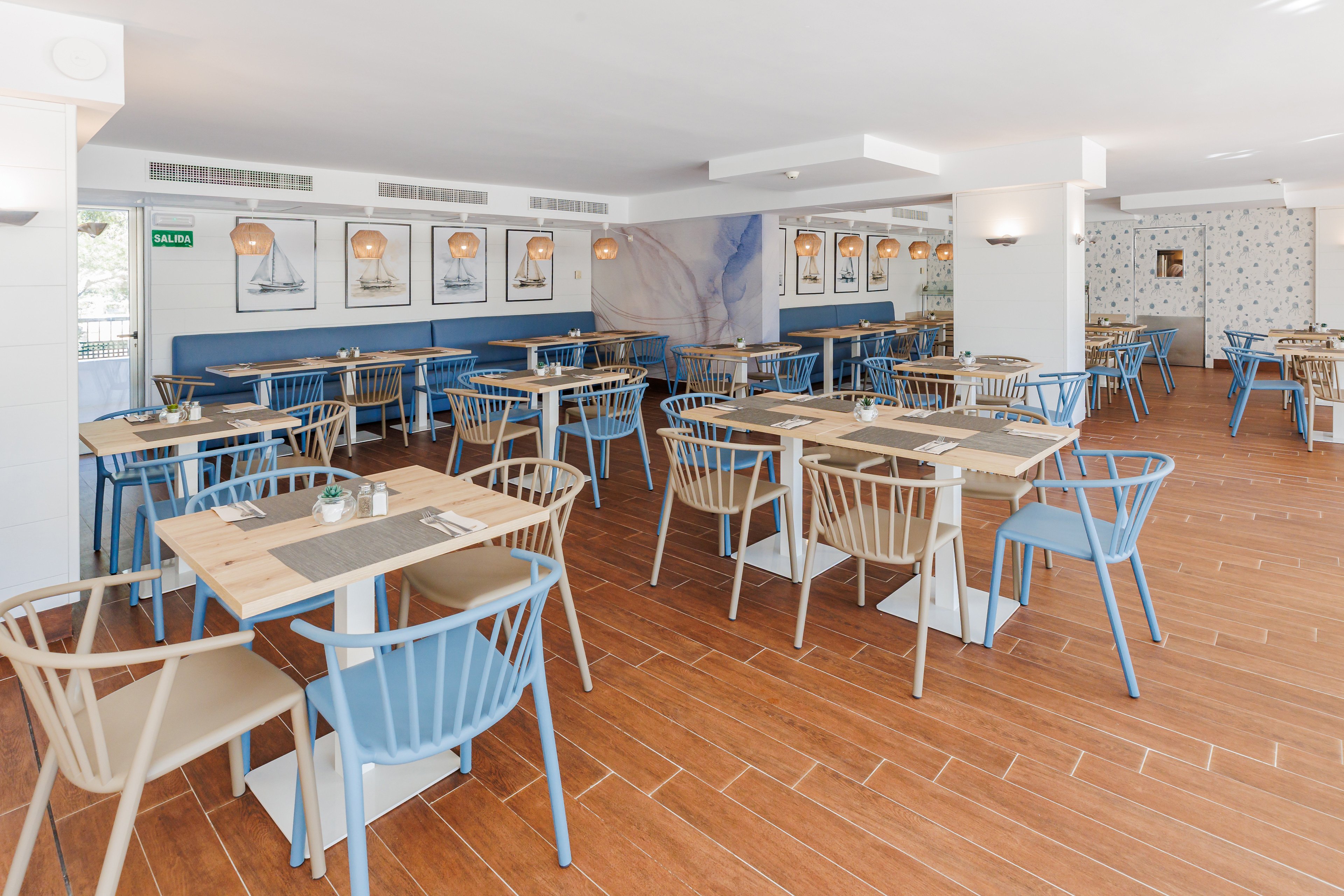 Spacious restaurant dining area featuring light blue and beige chairs around square tables. The setting is bright and airy with wooden flooring, nautical-themed wall art, and soft pendant lighting. The overall mood is relaxed and inviting, with a coastal-inspired palette and minimalist decor. No people are present, and the room appears ready for guests. The word 'SALIDA' is visible on a green exit sign.