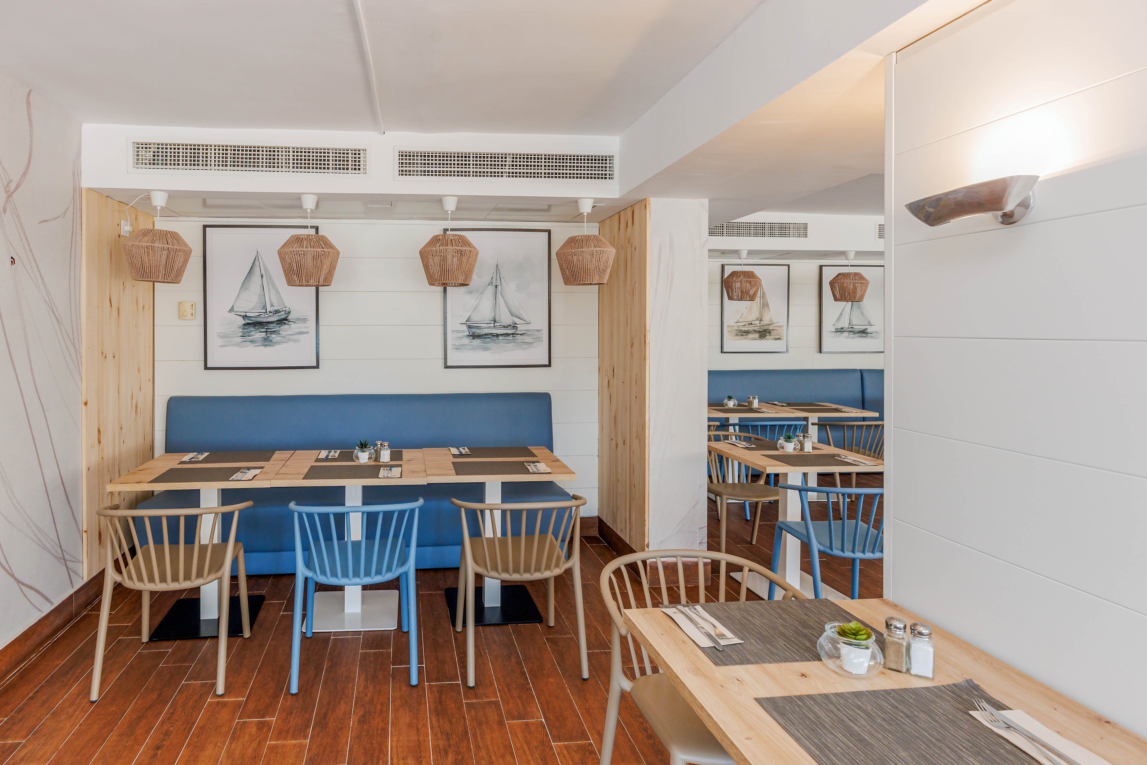 A stylish restaurant dining area features wooden chairs, blue bench seating, and nautical-themed wall art with sailboats. The setting is bright and inviting, with woven pendant lights and neatly set tables. The decor combines natural wood tones with white walls, creating a relaxed and contemporary atmosphere. Table settings include placemats, cutlery, and small potted plants, enhancing the welcoming vibe.