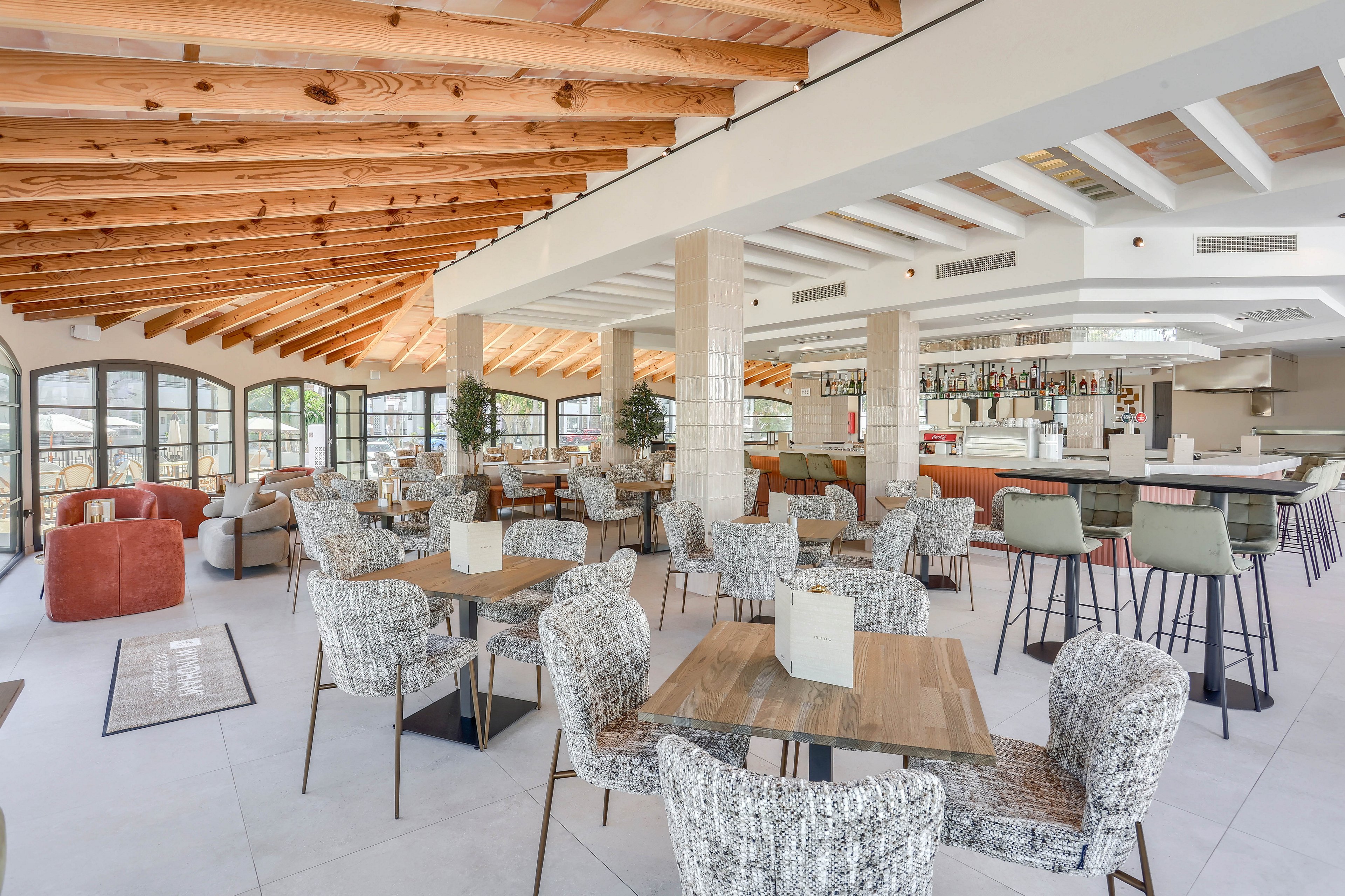 Spacious restaurant dining area featuring contemporary furniture and a stylish bar. The setting includes patterned chairs, wooden tables, and a ceiling with exposed wooden beams, creating a bright and inviting atmosphere. Large windows allow natural light to fill the space, while neutral tones and soft accents enhance the modern aesthetic. No visible text or numeric values are present in the image.