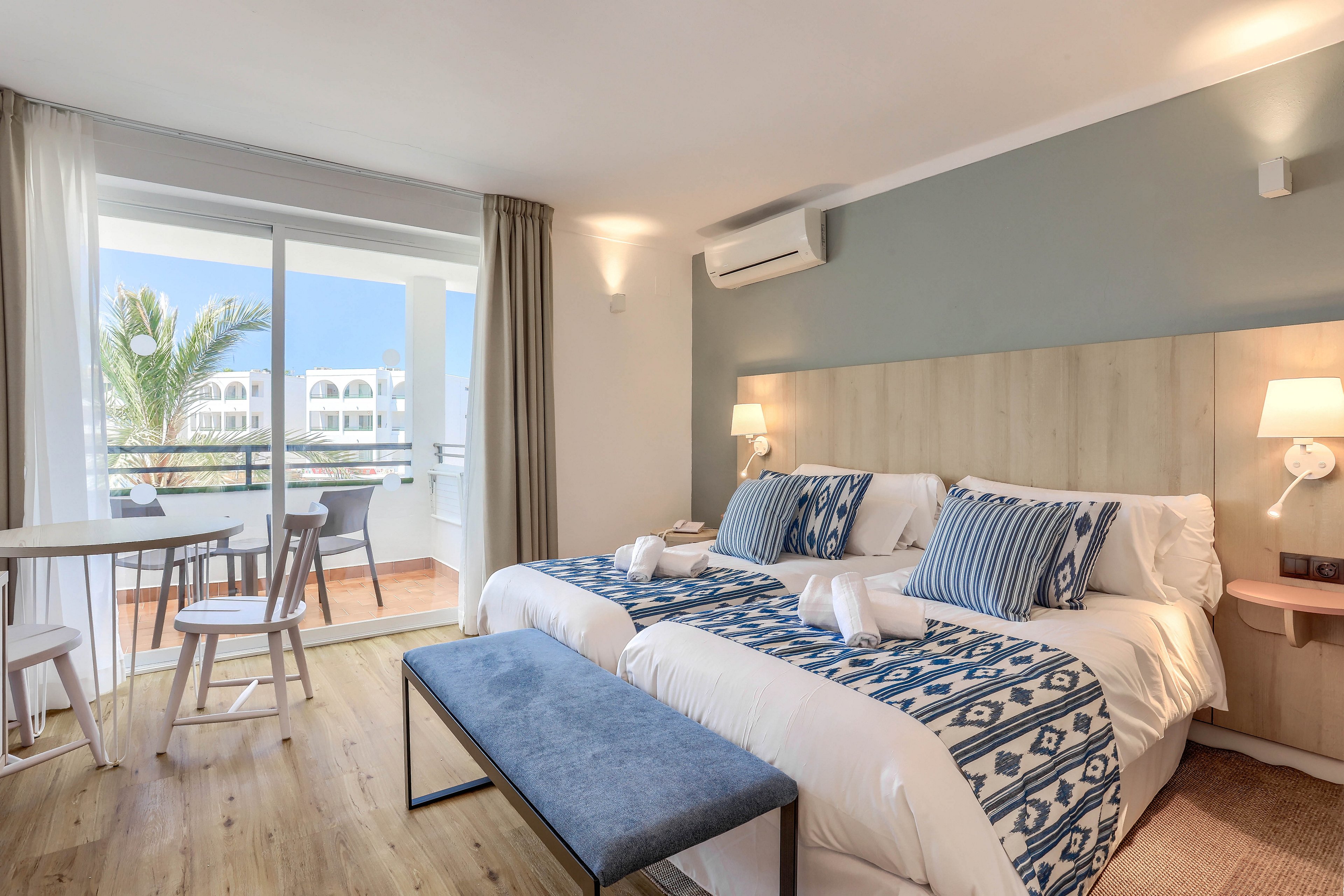A contemporary hotel room features two neatly made twin beds with patterned blue and white bedding. The space includes a small dining table and chairs near a large sliding glass door that opens to a sunny balcony with palm tree views. Soft lighting, wooden floors, and neutral tones create a calm and inviting atmosphere. An air conditioning unit is visible above the beds, and towels are arranged on the beds.