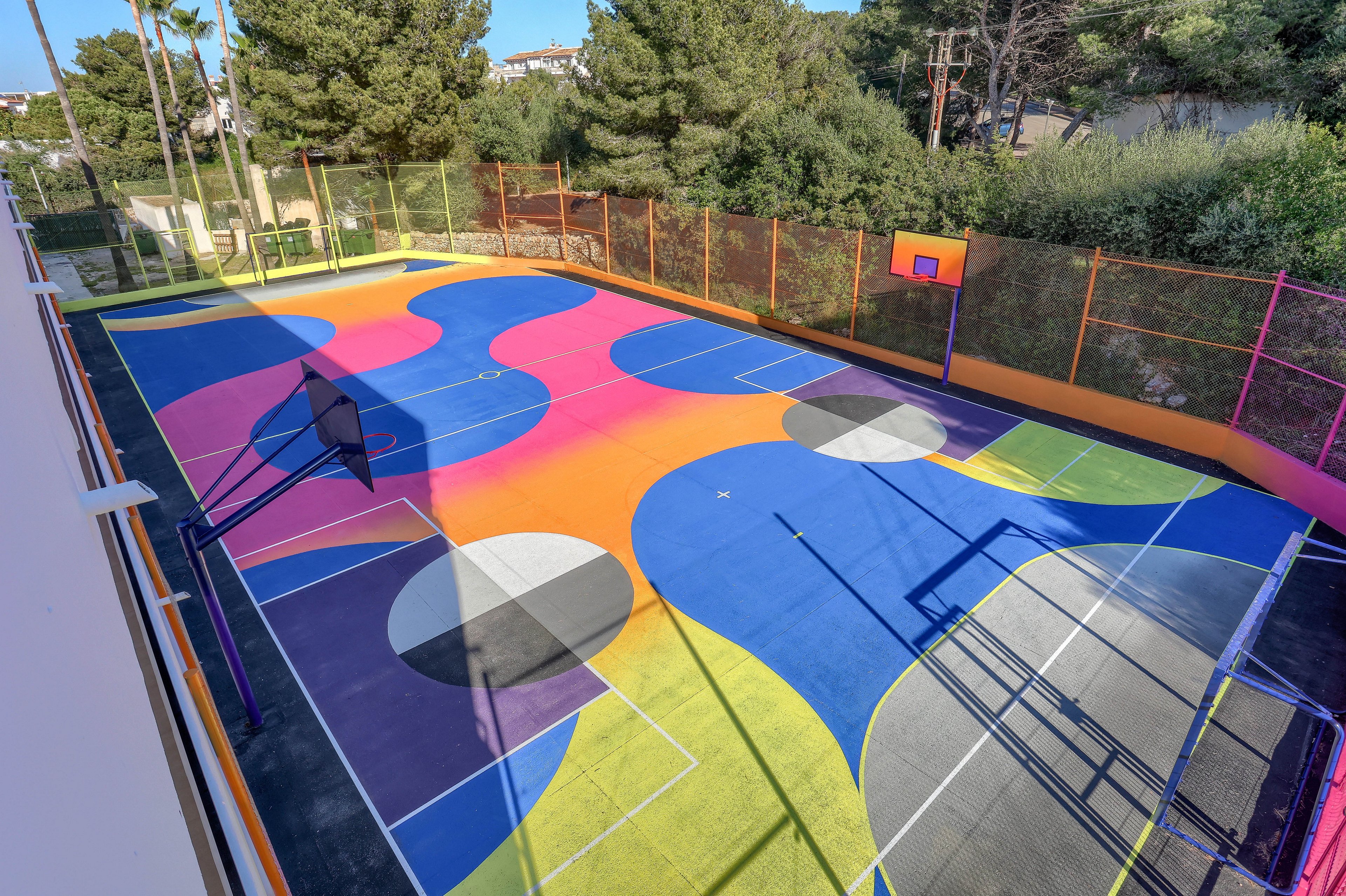 The main subject is a vibrant outdoor basketball court featuring an abstract, multicolored pattern with flowing shapes in blue, orange, pink, yellow, and gray. The setting is surrounded by trees and fencing, with a basketball hoop visible at one end. The court is empty, highlighting the artistic visuals and geometric lines. No visible text or numbers are present in the image.
