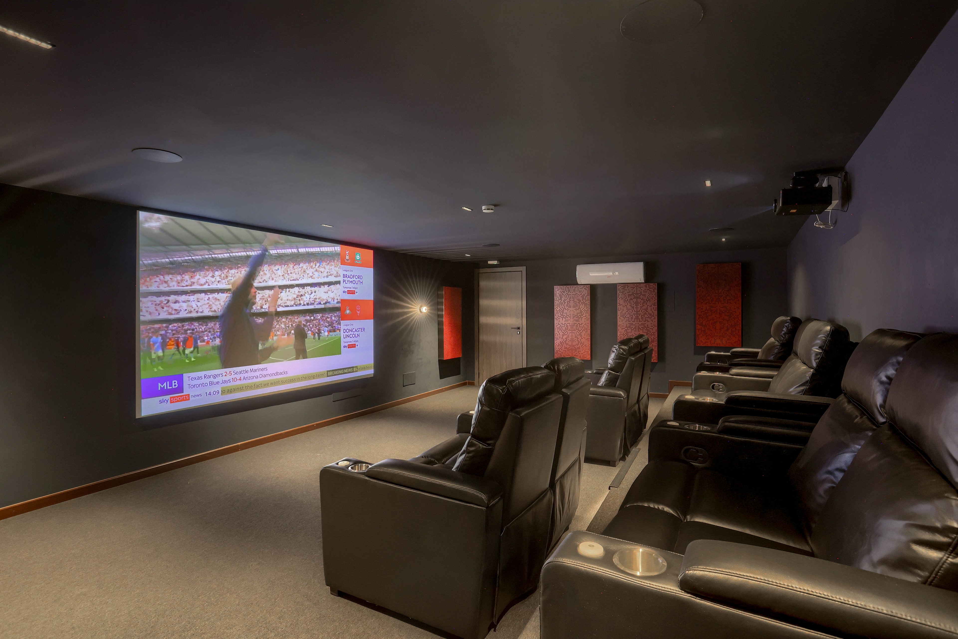 A contemporary home theater features several plush leather recliners arranged in rows facing a large wall-mounted projector screen. The screen displays a live sports event with visible text and graphics, creating an immersive viewing experience. The room is dimly lit with dark walls and acoustic panels, enhancing the cinematic atmosphere. No people are present, and the setting emphasizes comfort and entertainment.