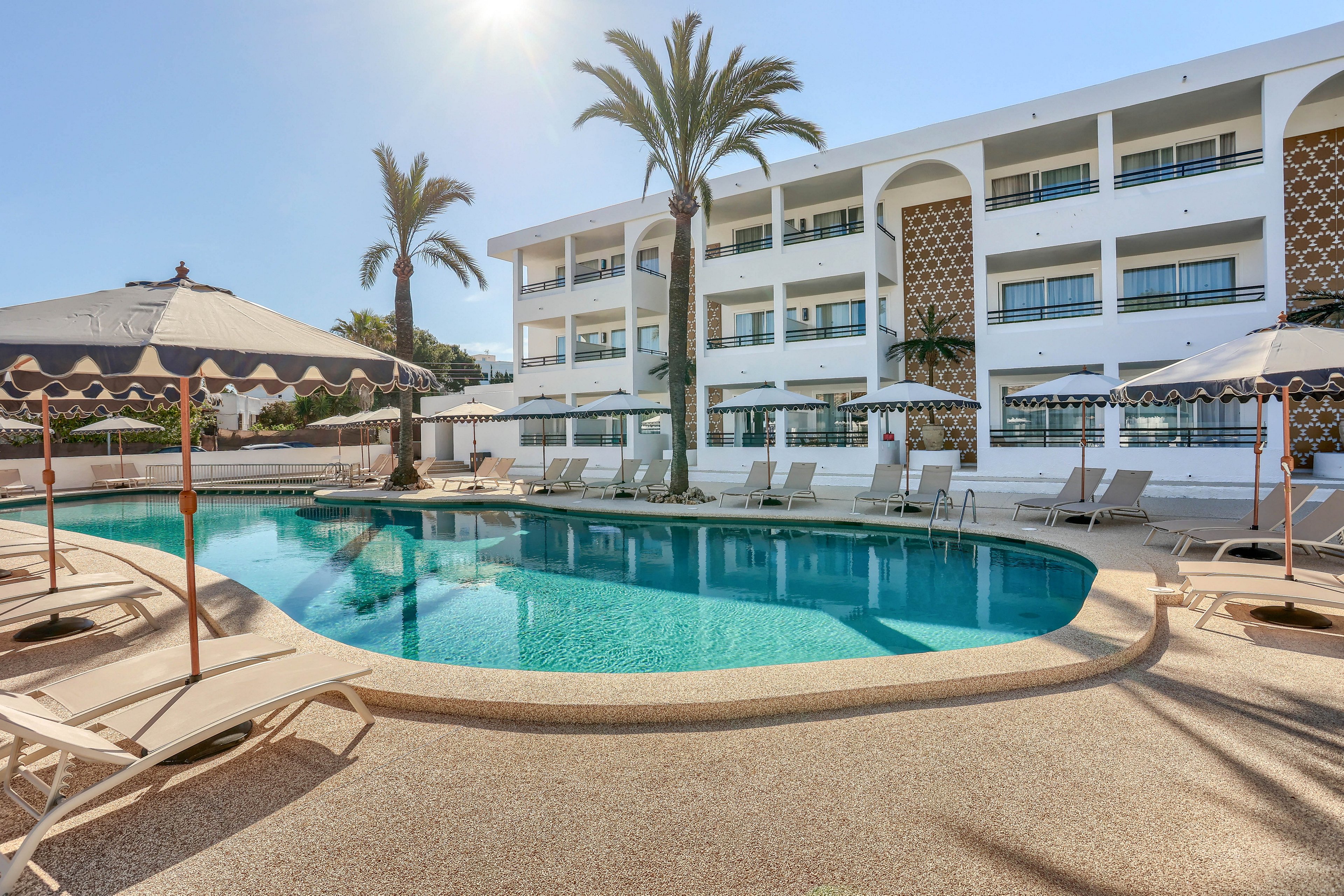 The image features a contemporary hotel pool area surrounded by sun loungers and large umbrellas. The setting is outdoors, with palm trees adding a tropical touch and a white multi-story hotel building in the background. The pool water is clear and inviting, reflecting the sunny sky and creating a relaxed atmosphere. The overall mood is tranquil and leisurely, ideal for vacation or relaxation.