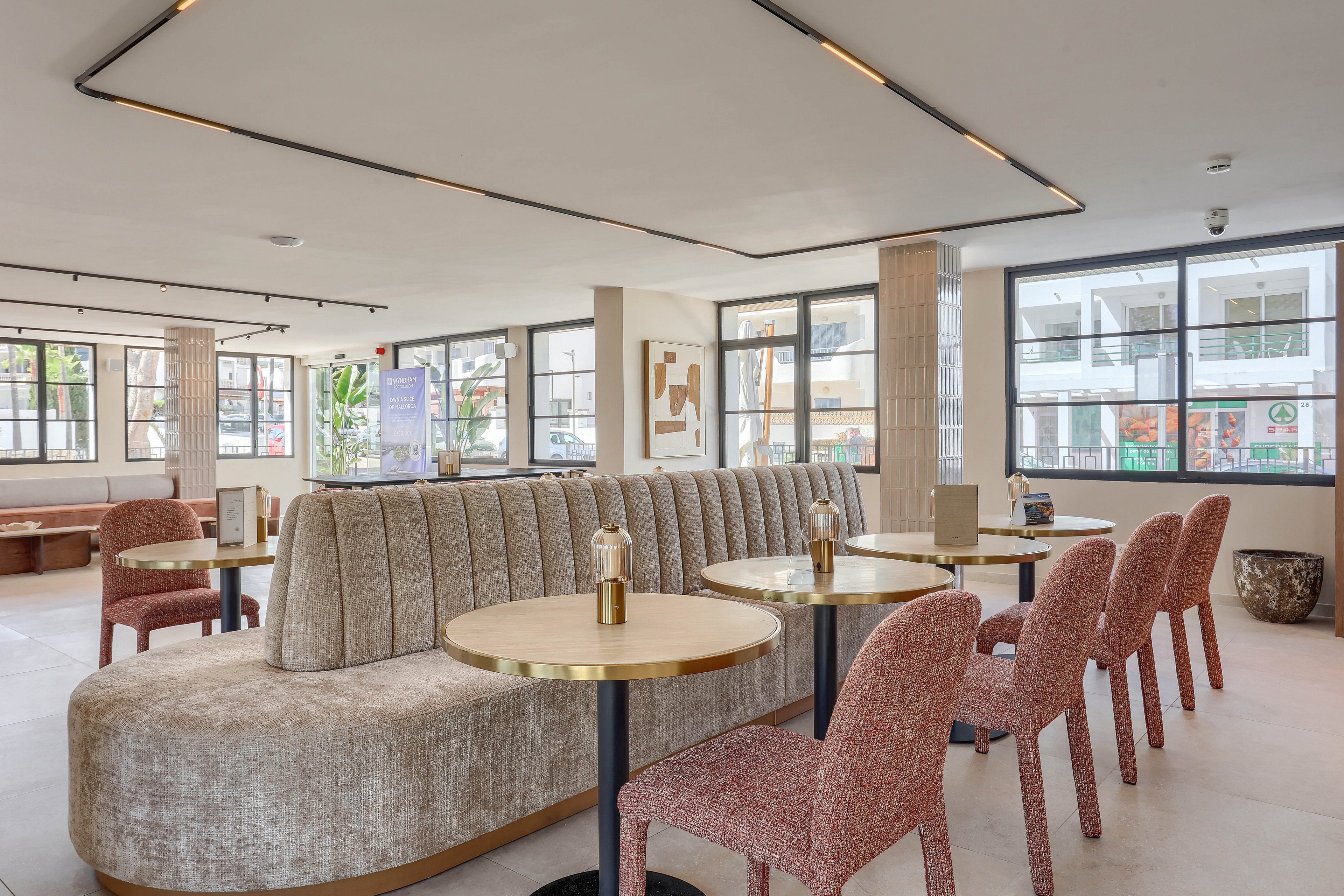 A contemporary lounge space features curved beige upholstered benches and round tables paired with textured pink chairs. Large windows allow natural light to fill the open, airy room, highlighting minimalist decor and neutral tones. The setting suggests a relaxed, inviting atmosphere suitable for socializing or casual meetings.