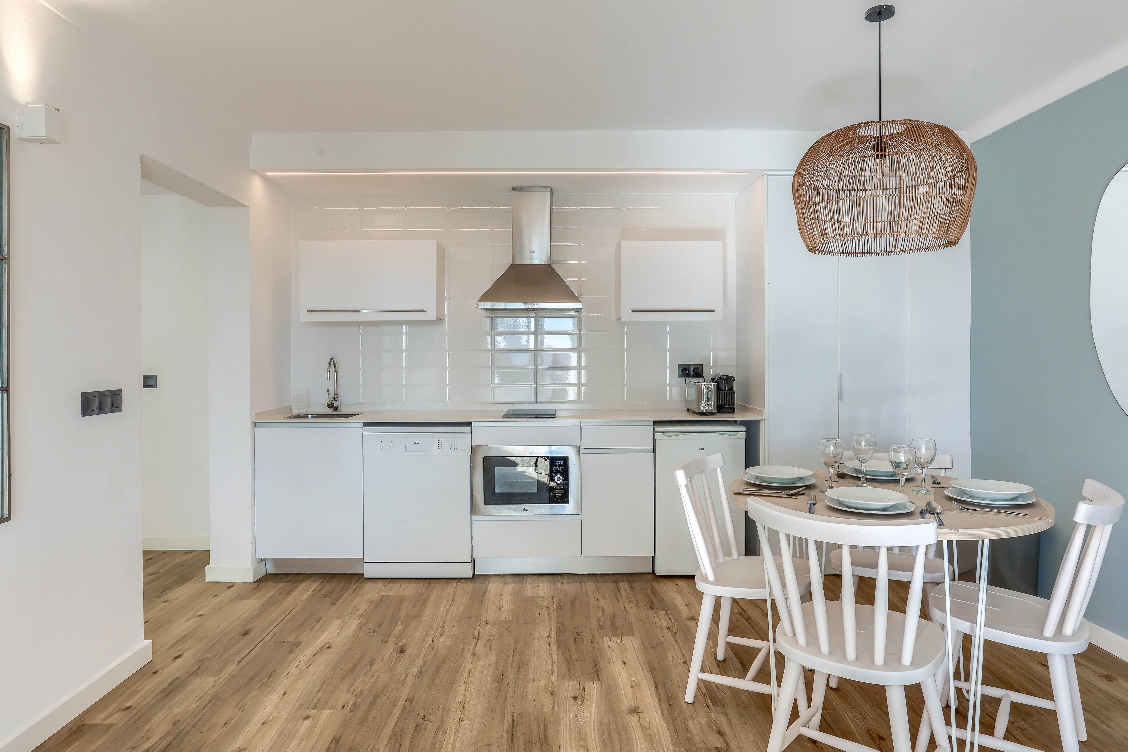 A contemporary kitchen features sleek white cabinetry, integrated appliances, and a stainless steel range hood. The space includes a round dining table set for four, with white chairs and a woven pendant light overhead. Light wood flooring and a soft blue accent wall create a calm, inviting atmosphere.