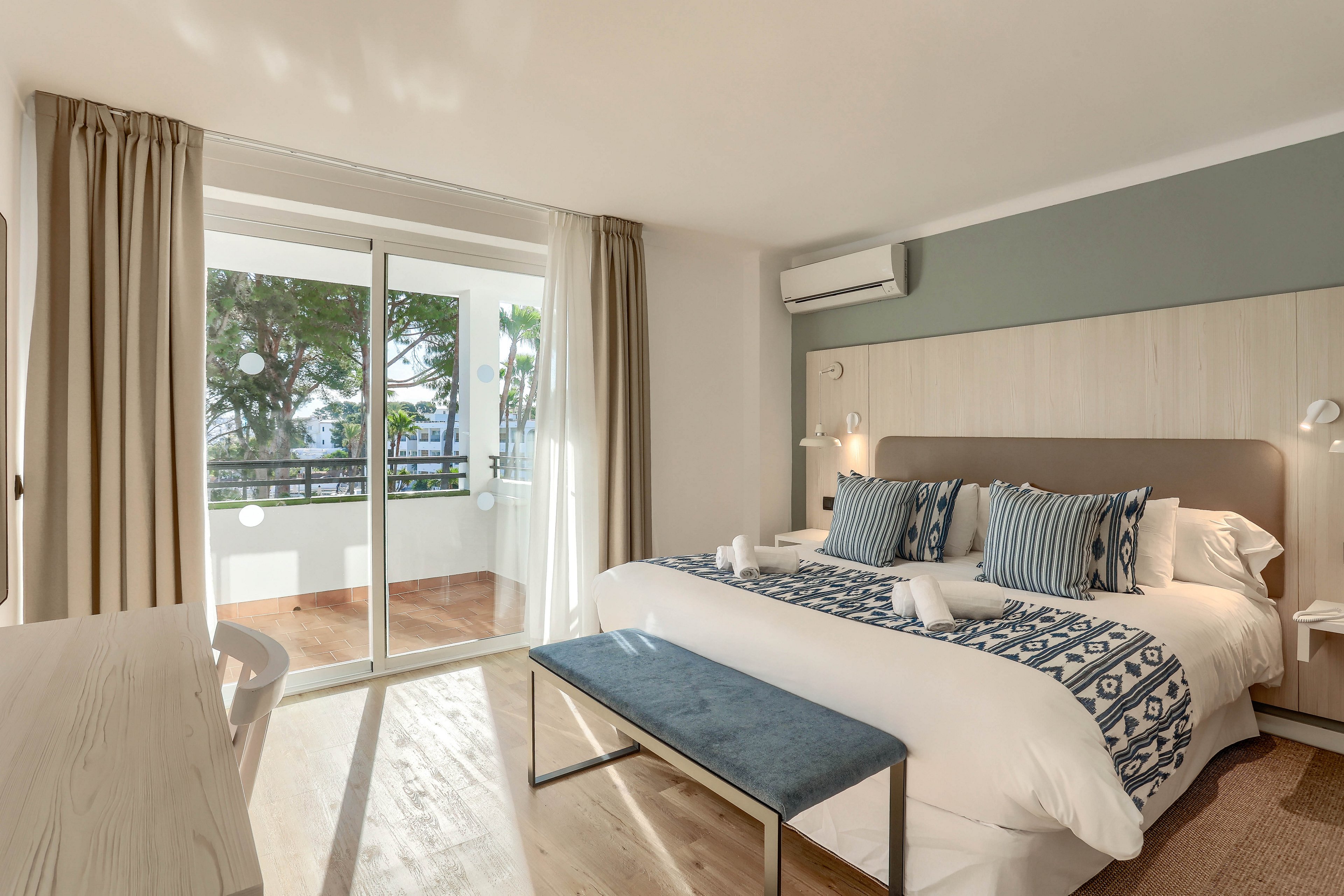 A contemporary hotel room features a large bed with patterned pillows and a bench at the foot. The space is bright, with sunlight streaming through glass doors that open to a balcony overlooking greenery. Neutral tones and minimalist decor create a calm, inviting atmosphere. No visible text or numeric values are present in the image.