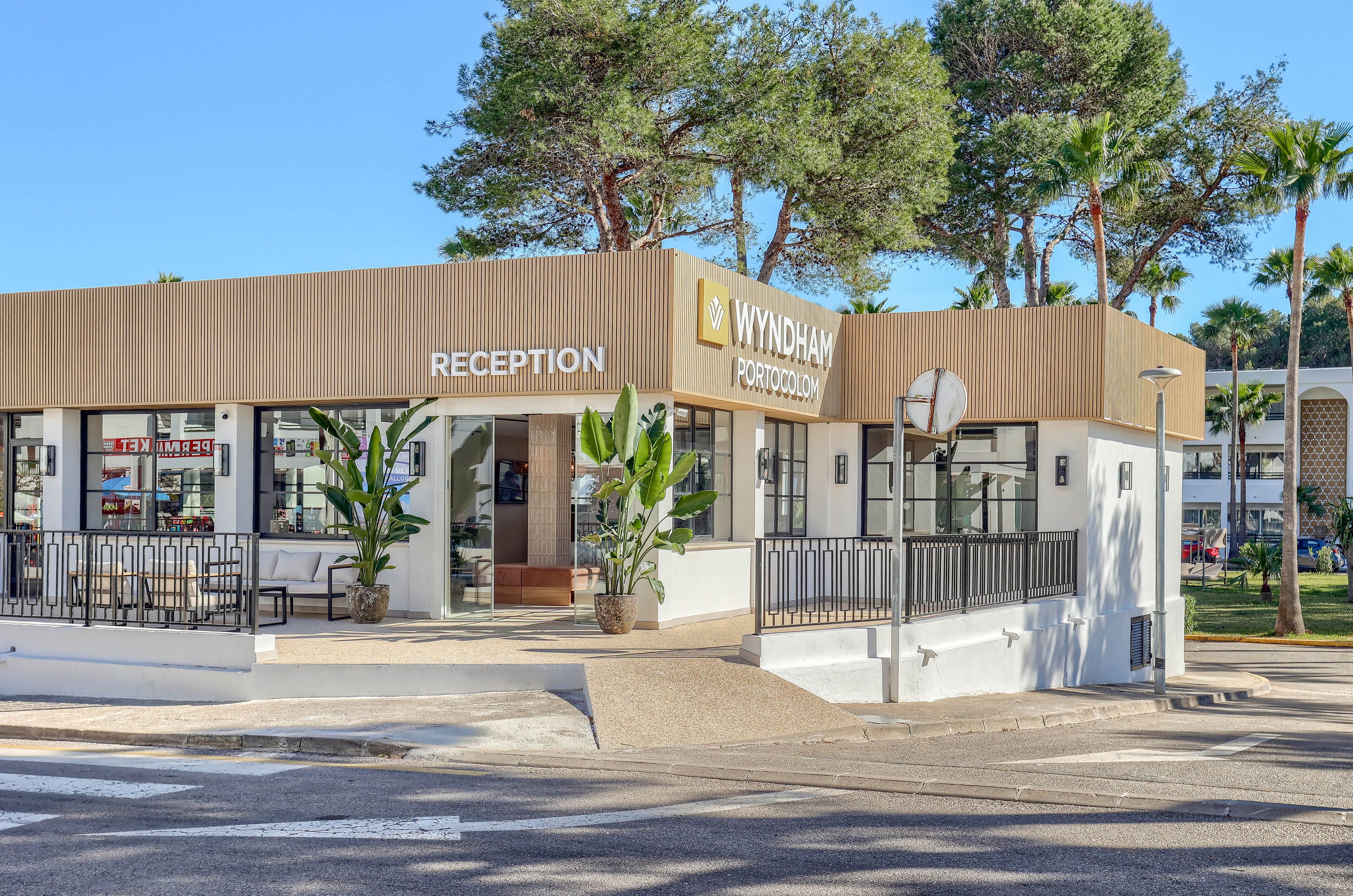 The image shows the exterior of a Wyndham hotel reception building, clearly marked with the 'WYNDHAM' and 'RECEPTION' signs. The setting features a modern architectural style with large windows, potted plants, and a clean, neutral color palette. The surrounding area includes palm trees and a sunny outdoor environment, suggesting a resort or vacation destination. No people are visible in the scene.