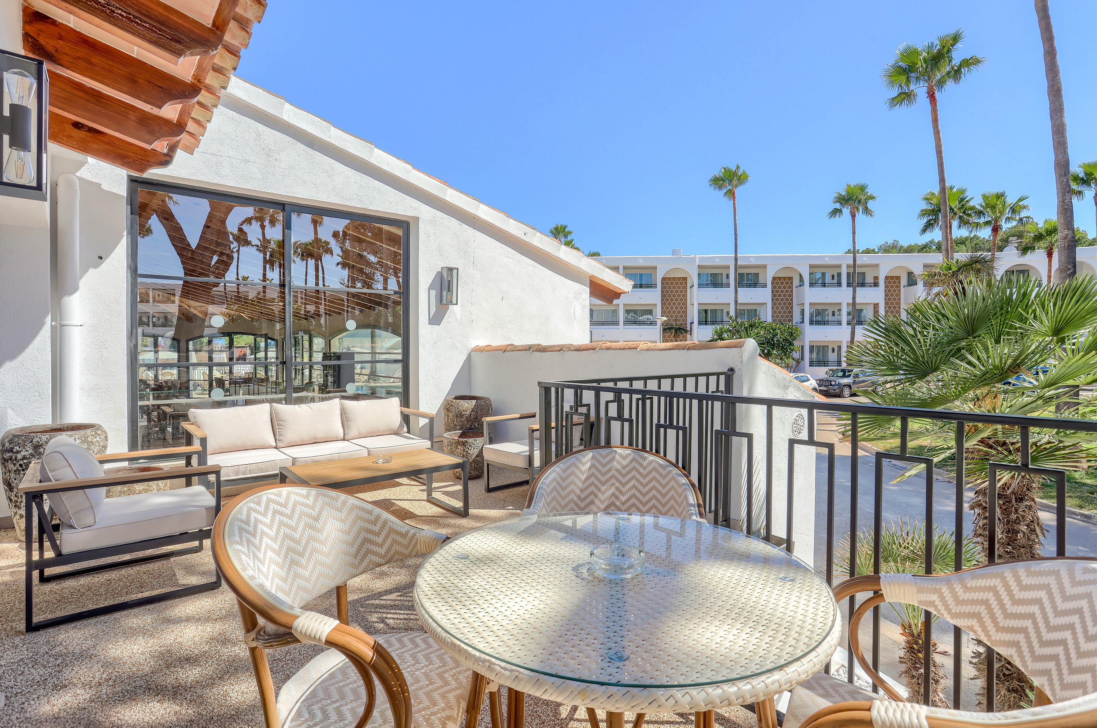 A bright, sunlit balcony features a round glass table and four woven chairs, overlooking a resort courtyard with palm trees. The setting includes a comfortable outdoor sofa and modern architecture with white walls and large windows. The atmosphere is relaxed and inviting, ideal for leisure or social gatherings. The clear blue sky and lush greenery enhance the tranquil, vacation-like mood.