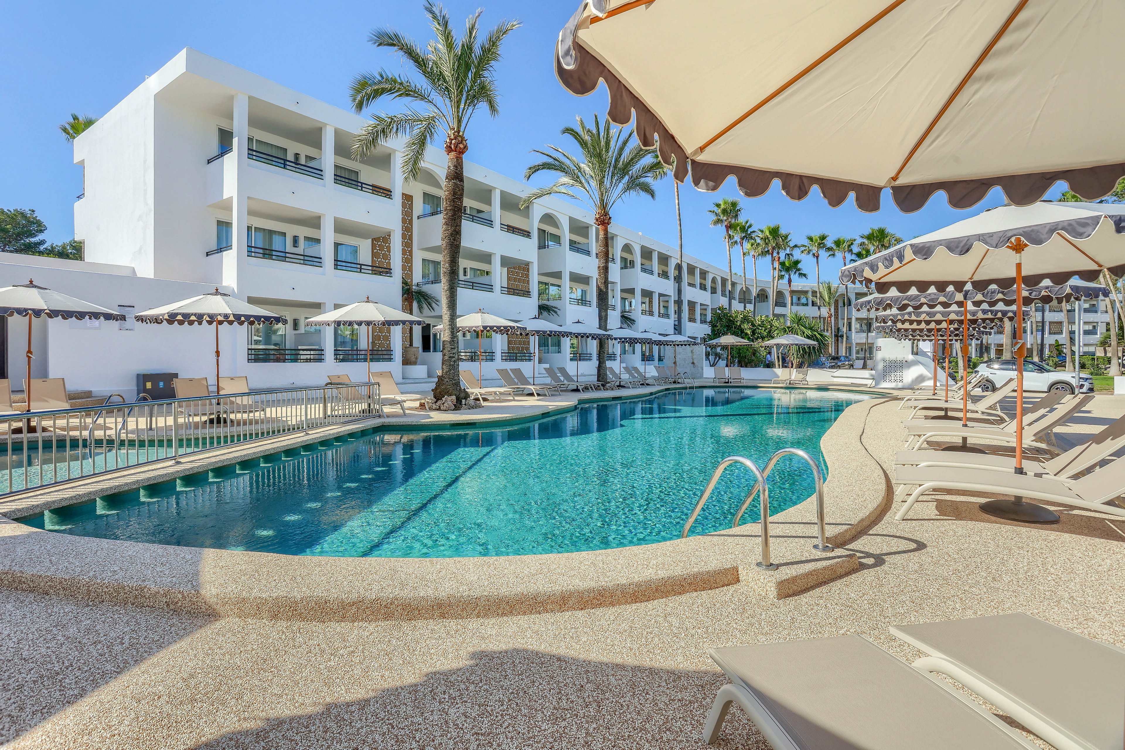 A contemporary hotel features a spacious outdoor swimming pool surrounded by beige sun loungers and large parasols. The setting is bright and sunny, with palm trees lining the poolside and white buildings in the background. The scene evokes a relaxed vacation mood, with clear blue water and minimalist architecture. No visible text or numbers are present in the image.