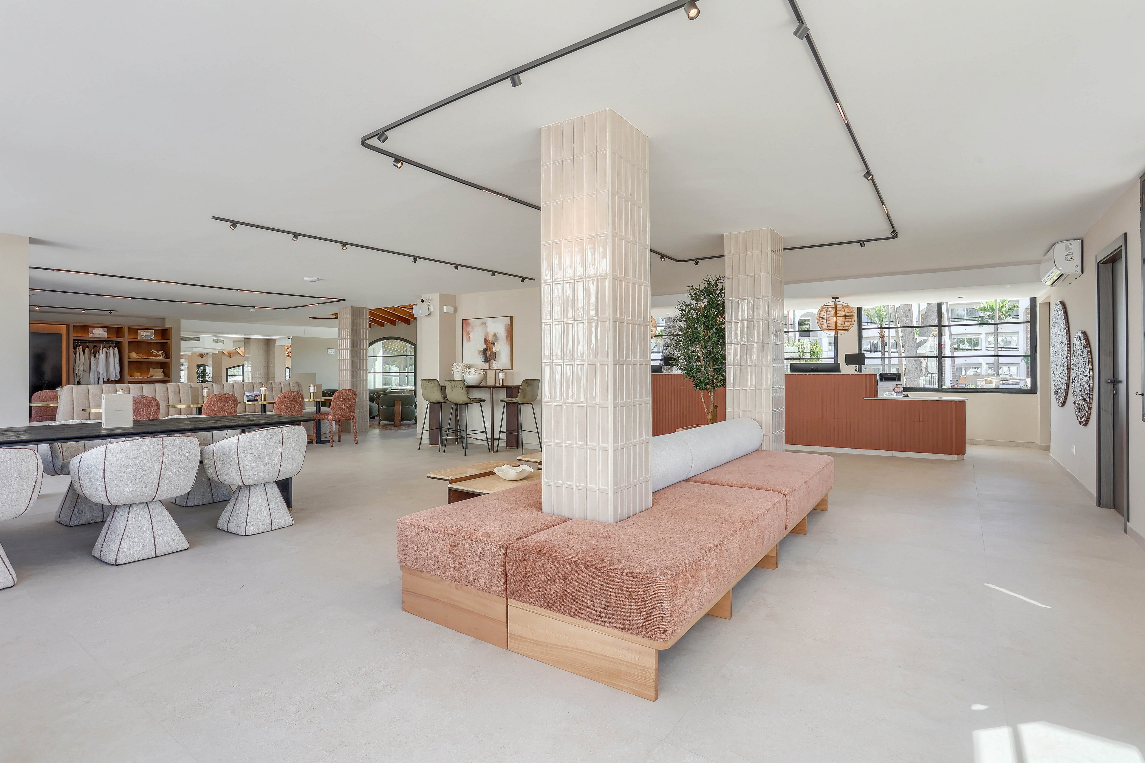 Spacious contemporary lounge area featuring minimalist furniture and neutral tones. The setting includes a central seating bench, dining table with white cushioned chairs, and large windows allowing natural light. Decorative elements and plants enhance the airy, welcoming atmosphere. The space is uncluttered, with clean lines and soft textures throughout.