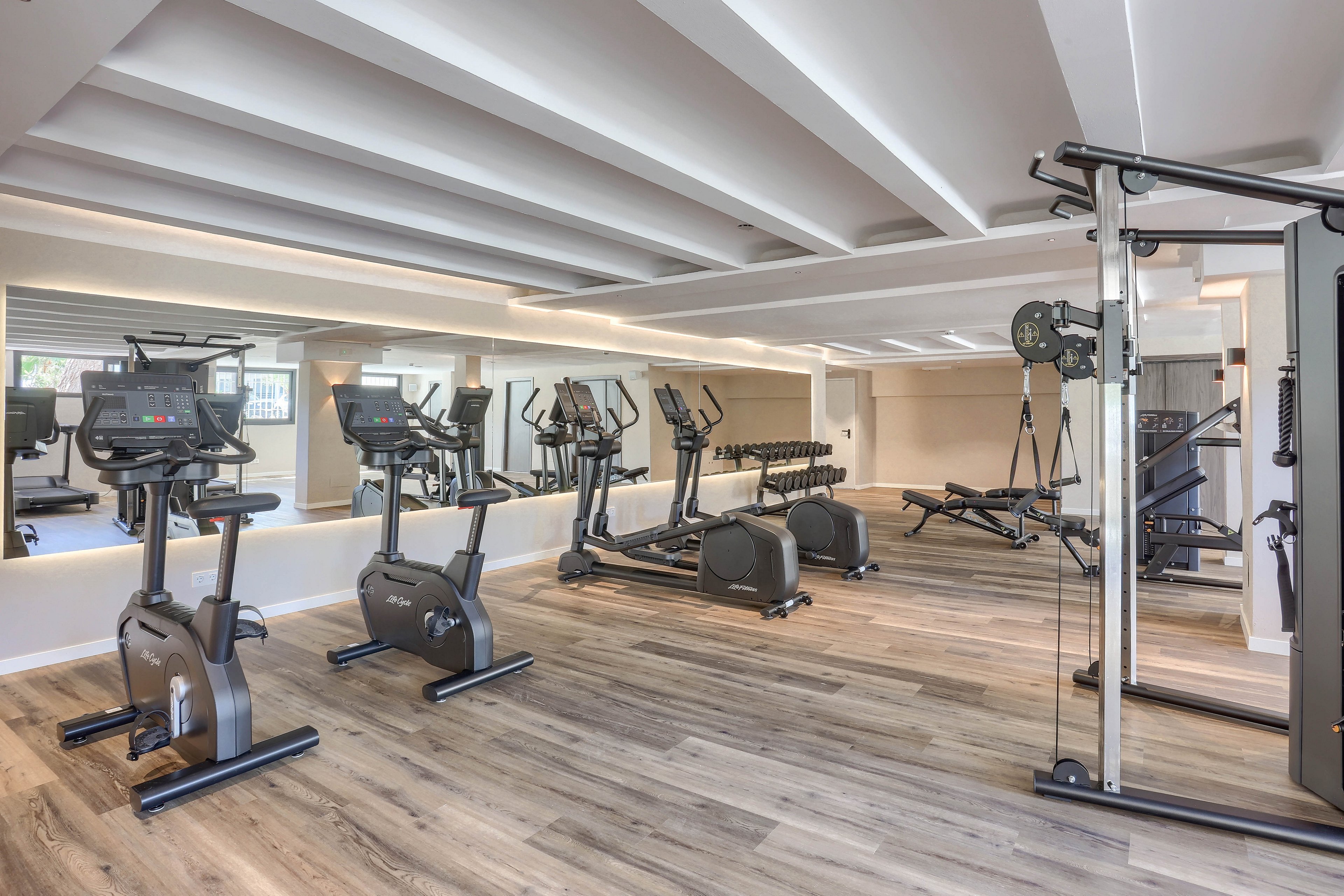 A spacious, contemporary gym features multiple exercise machines including treadmills, stationary bikes, and a cable weight system. The setting is bright with natural light, mirrored walls, and wood-style flooring. Dumbbells are neatly arranged on racks, and the overall palette is neutral with clean lines. No people are present, emphasizing the equipment and open layout.